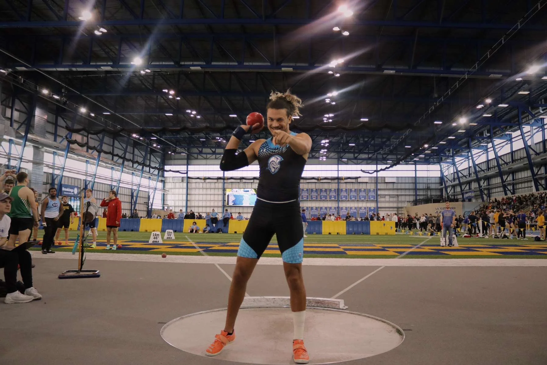 Treshawn preparing to throw at an indoor track and field event, with spectators and other athletes in the background.