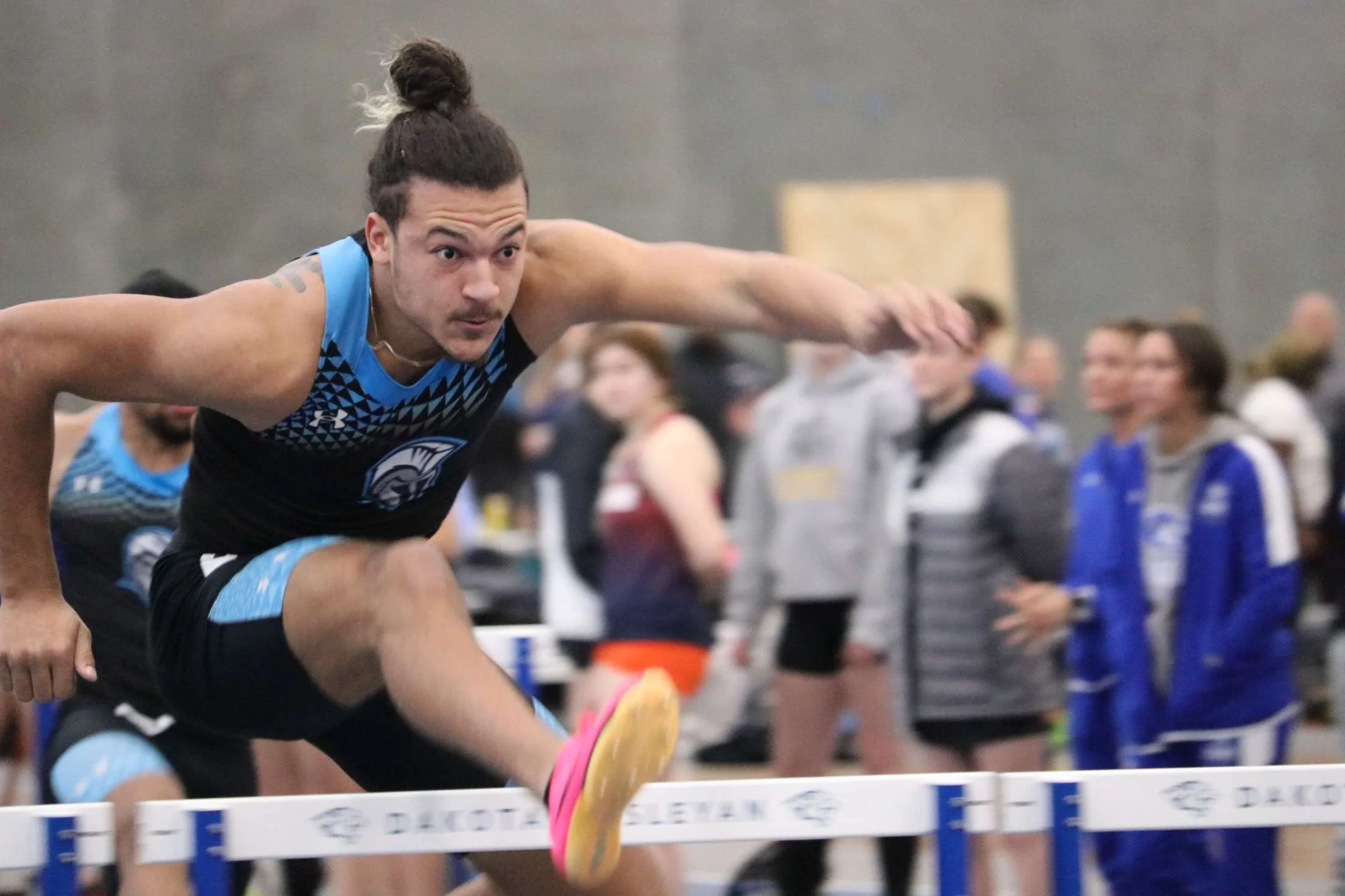 A male athlete with a man bun hairstyle wearing a blue and black athletic uniform, is mid-air jumping over a hurdle during an indoor track event, with an audience and other competitors in the background.