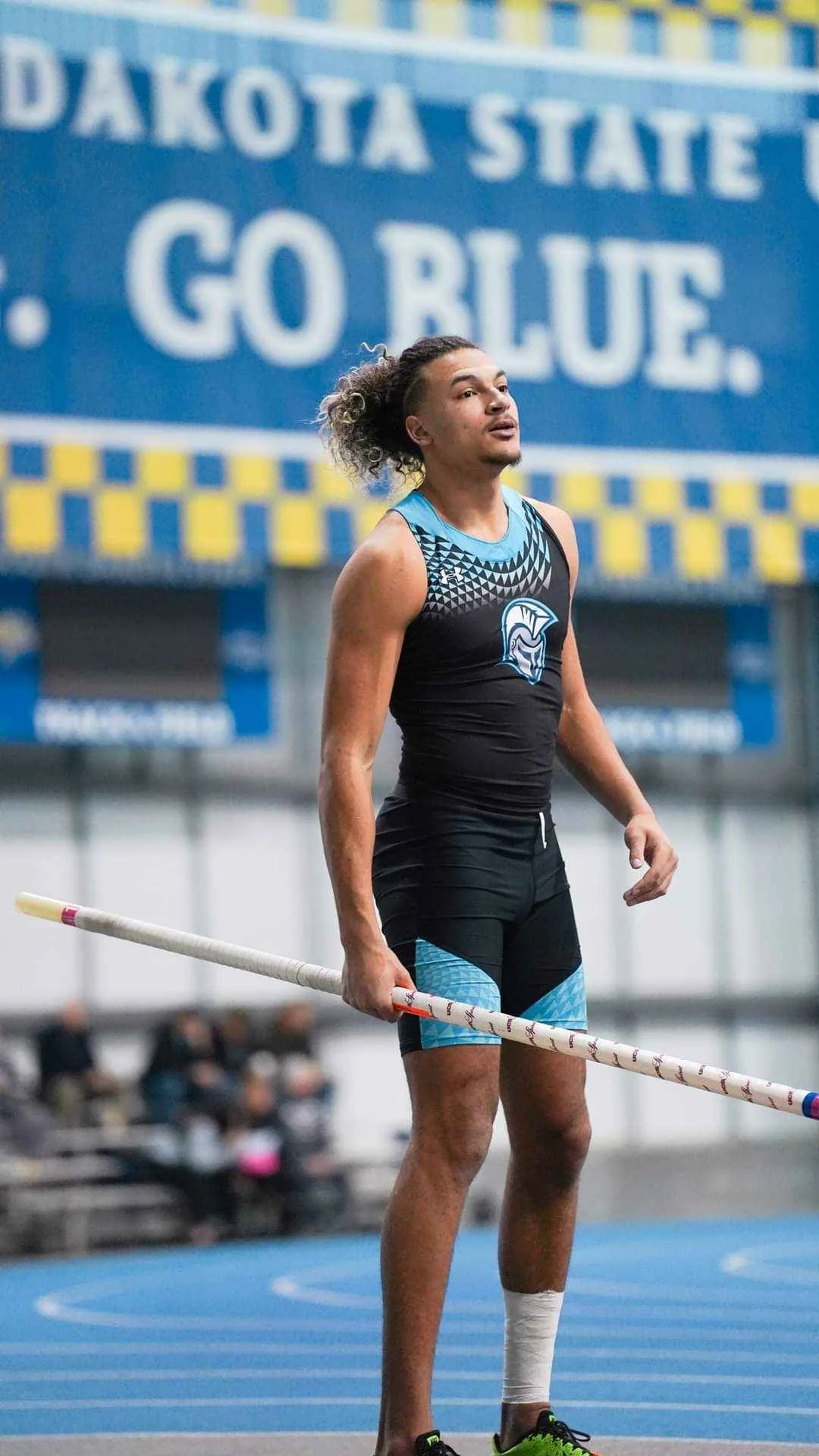 A male pole vaulter holding a pole, standing on an indoor track field, wearing a black and blue athletic uniform, with a Dakotа State sign in the background.