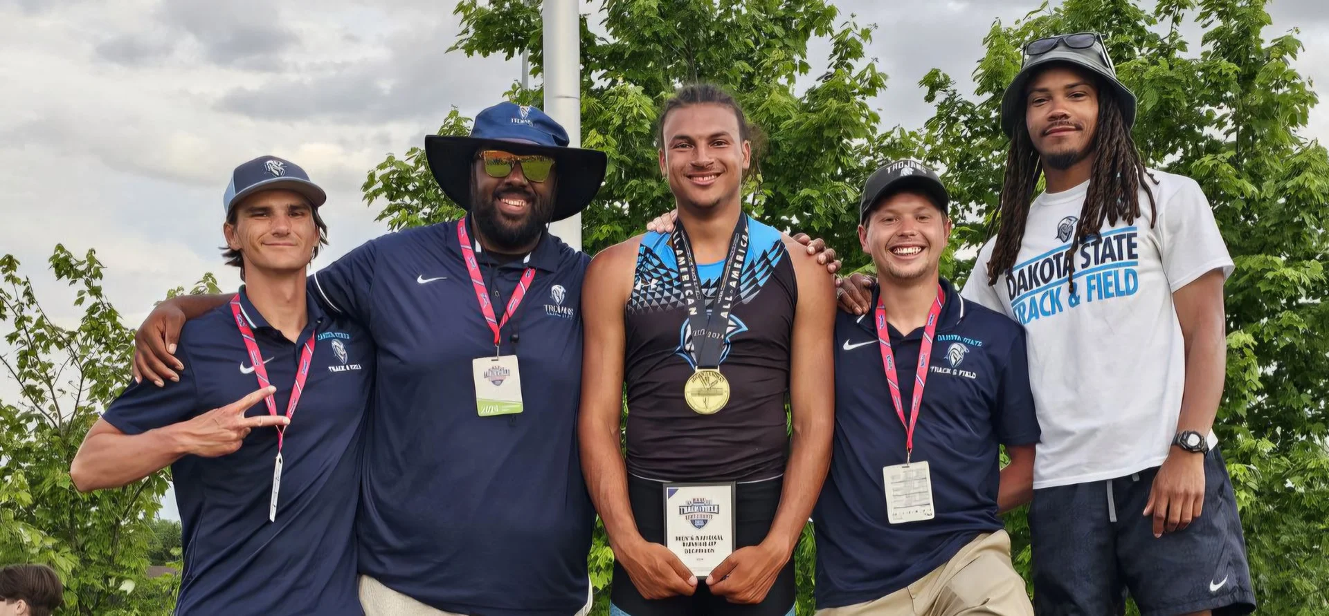 Group of five male athletes standing outdoors with medals, smiling, during a track and field event, with trees and cloudy sky in the background.