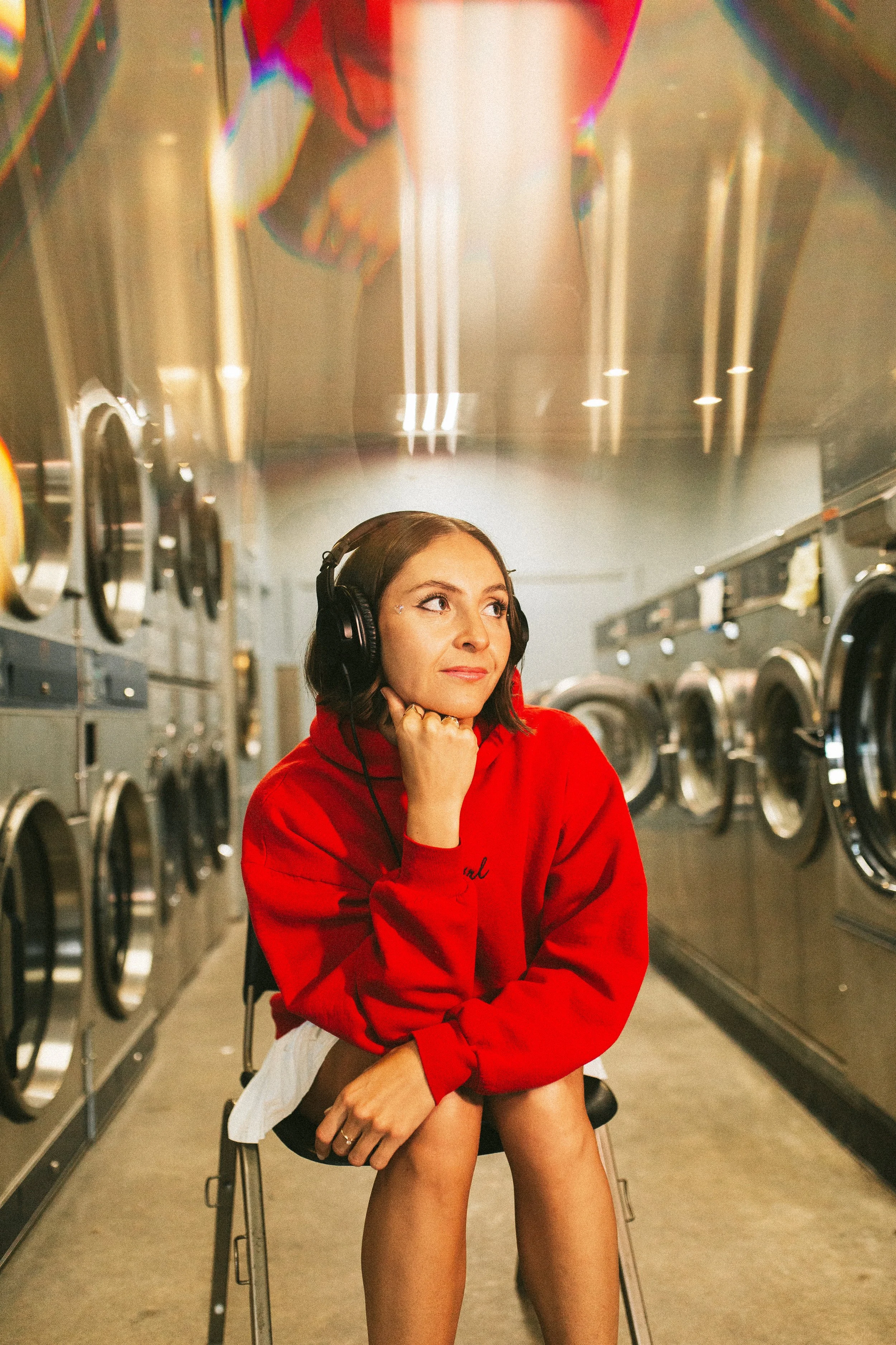Young woman sitting in a laundromat, wearing headphones and a red hoodie, with washing machines on both sides.