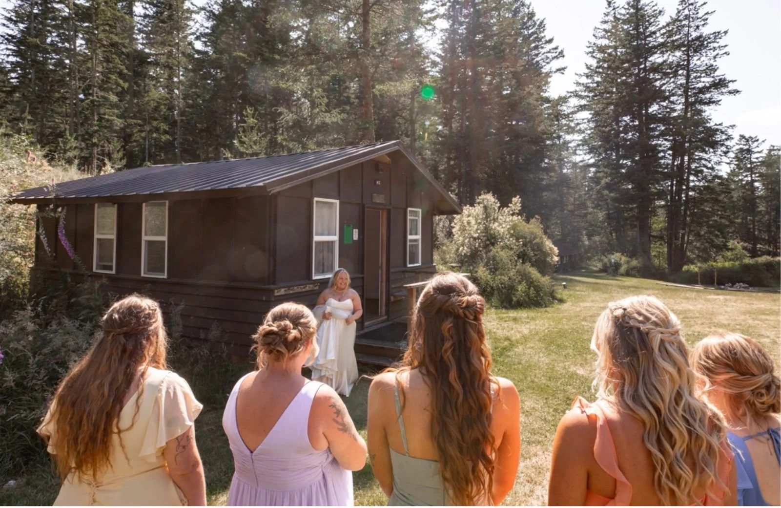 Women in light-colored dresses attending an outdoor wedding ceremony near a small dark wooden cabin, with a forested background on a sunny day.