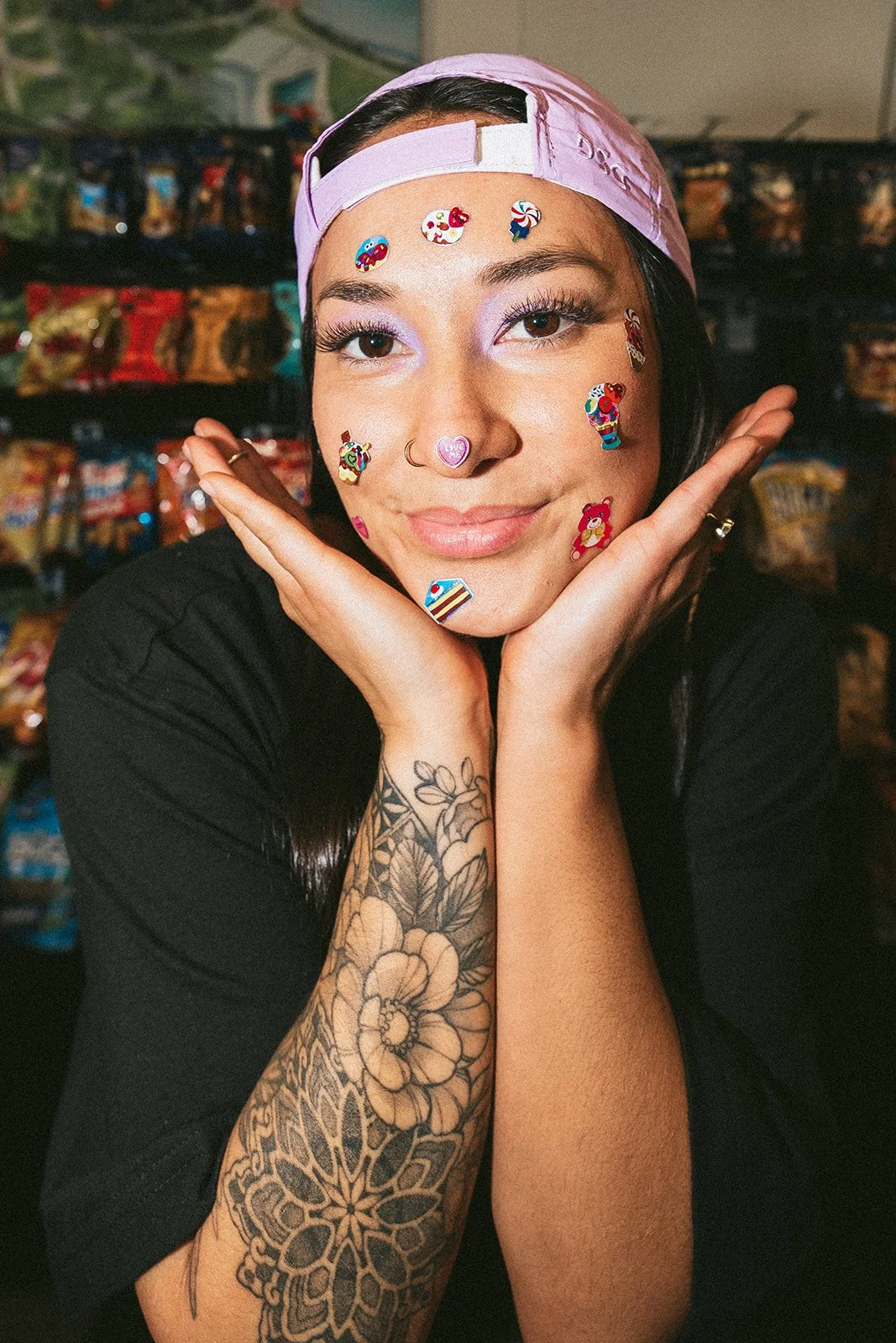 A woman with dark hair, wearing a light pink cap backward, decorated with colorful stickers of candy, bears, and desserts. She has purple eyeshadow, a nose ring, and a tattooed arm with flowers and mandala designs. She is resting her chin on her hands, smiling softly, in front of a background filled with snack bags.