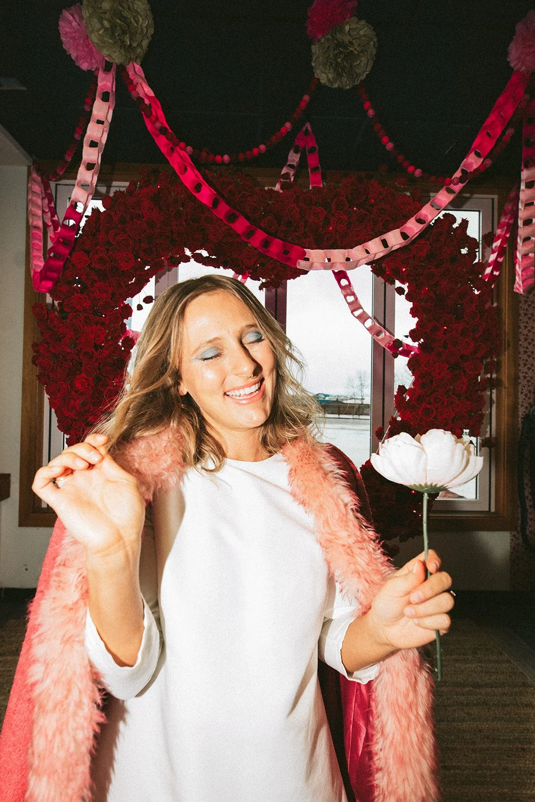 A woman smiling with closed eyes, holding a white flower, in front of a heart-shaped floral decoration made of red flowers, with pink and red streamers hanging from the ceiling.
