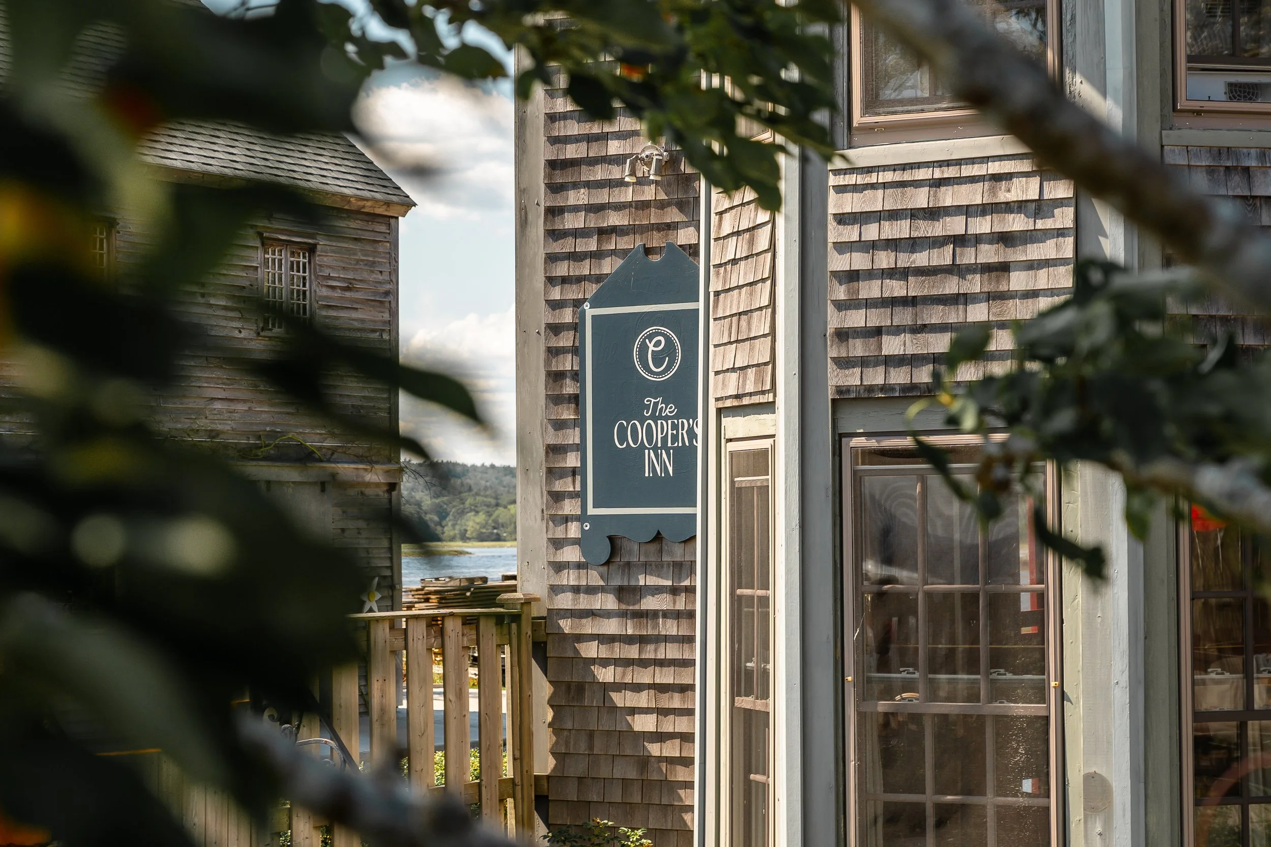 Outdoor view of a wooden building with a sign that reads 'The Cooper's Inn' on the wall, partially obscured by green leaves in the foreground. The building has windows and is situated near a body of water with a railing nearby.