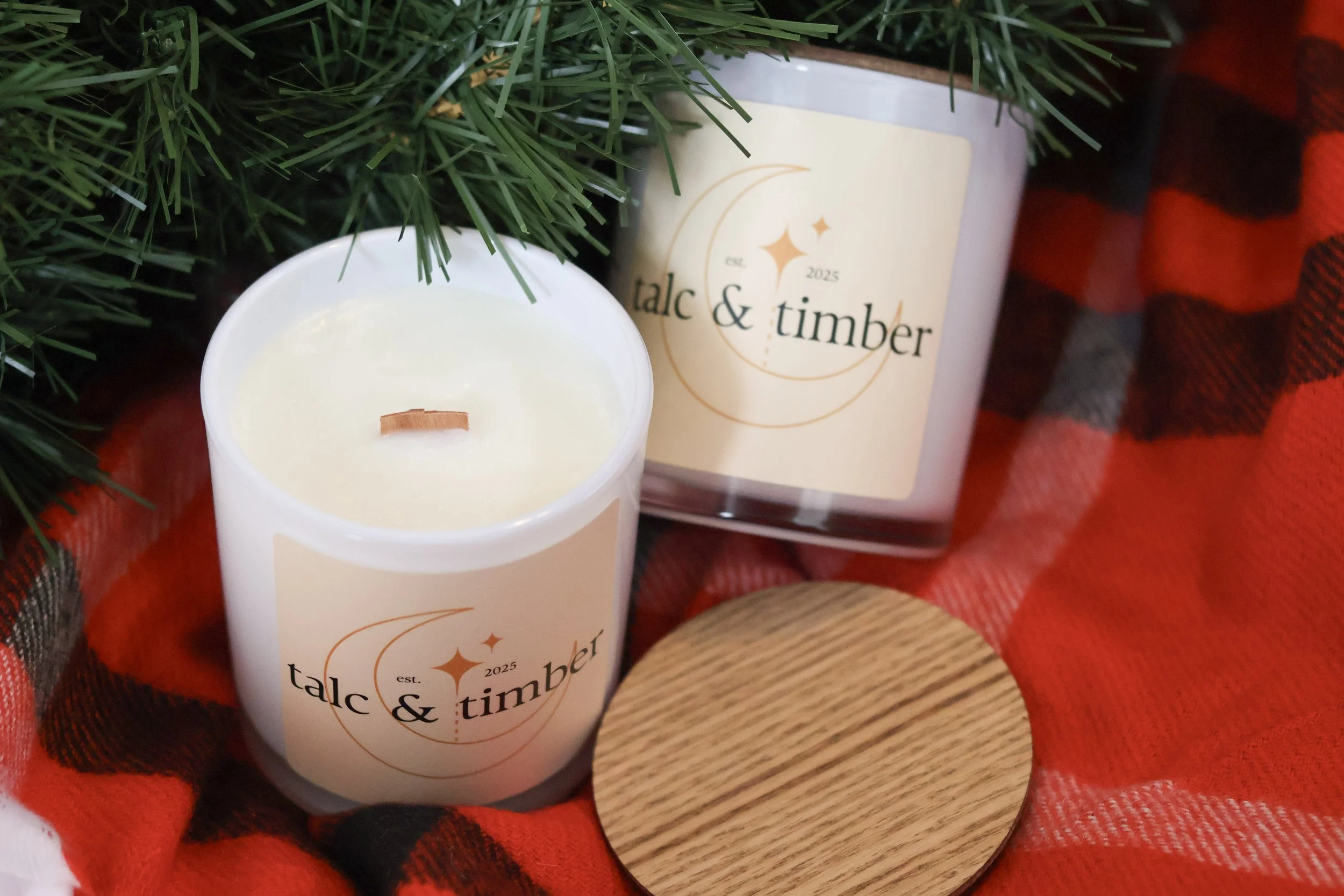 A lit white candle in a white jar with the logo "talc & timber" on it, placed on a red and black plaid fabric next to a green pine branch and a small round wooden lid.