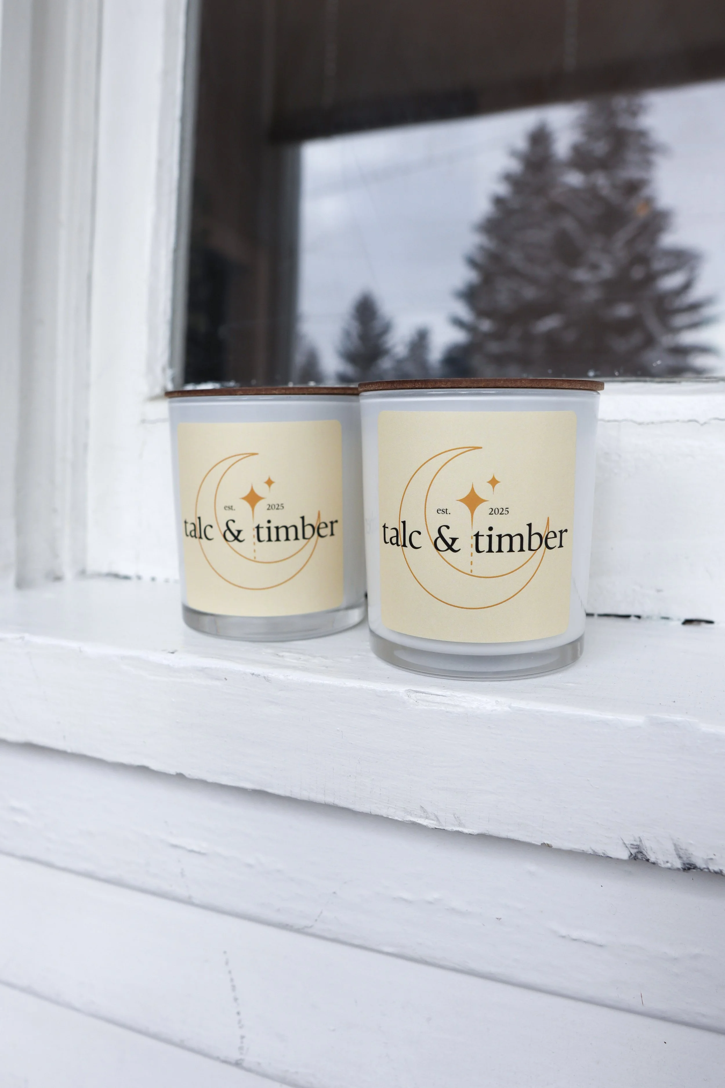Two candles with labels reading "talc & timber" placed on a white window ledge outside, with a window and trees in the background.
