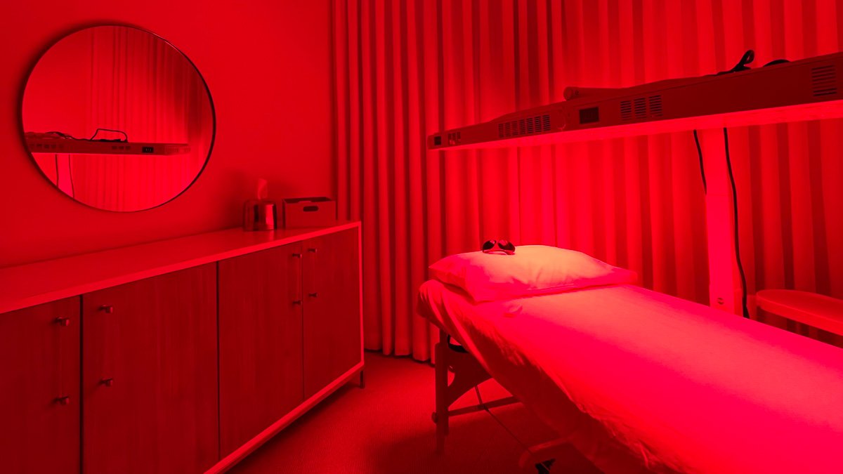 A medical or therapy room illuminated with red lighting, featuring a bed with a pillow, a small table with a device or object, a cabinet, a mirror on the wall, and curtains in the background.