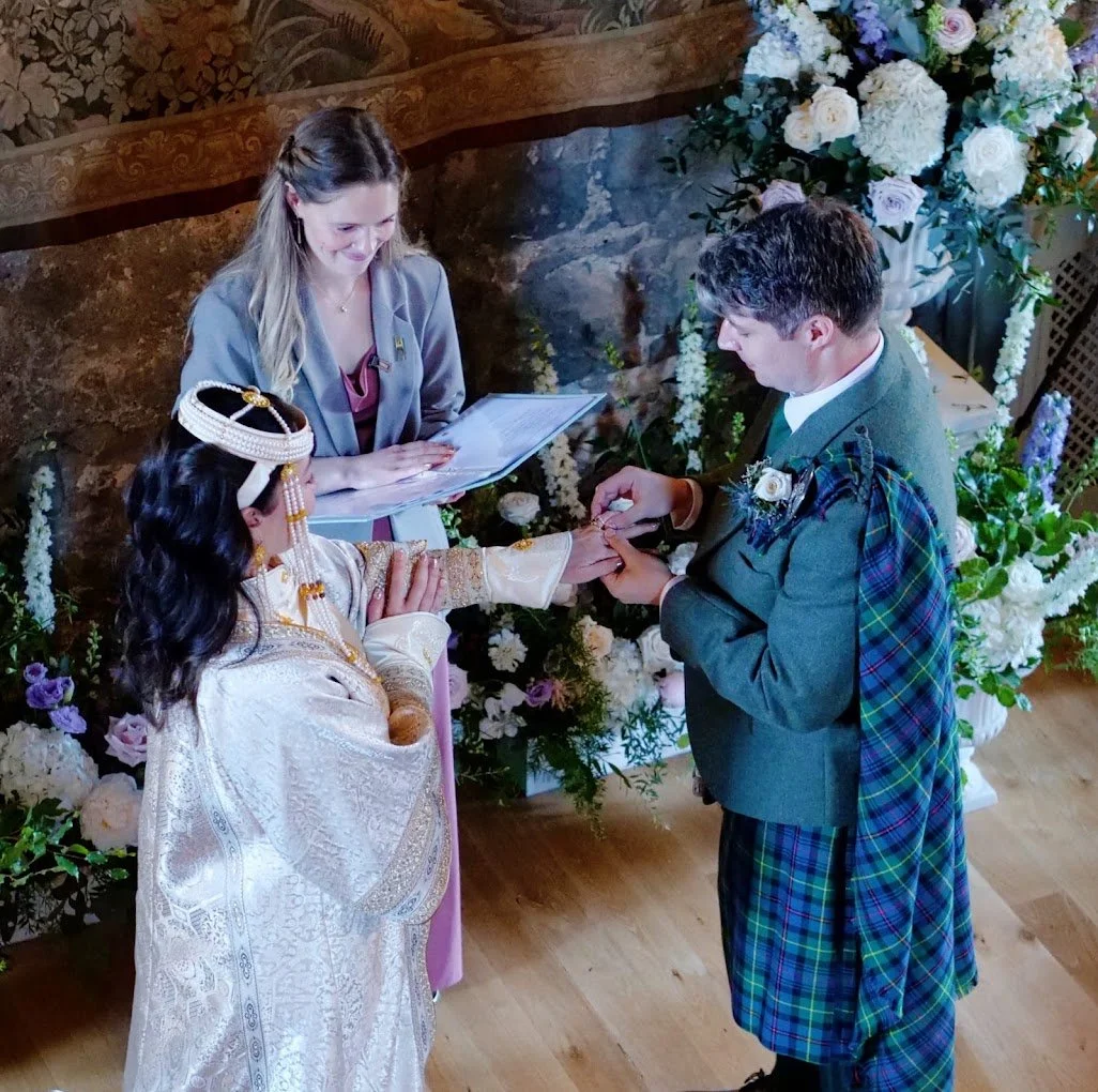 C and E, one of the Sunny Celebrant's couples, are exchanging rings. C wears a traditional tartan kilt, and E wears traditional Mongolian dress. Claire, holding a script, is standing in front of them smiling, next to some purple flowers.