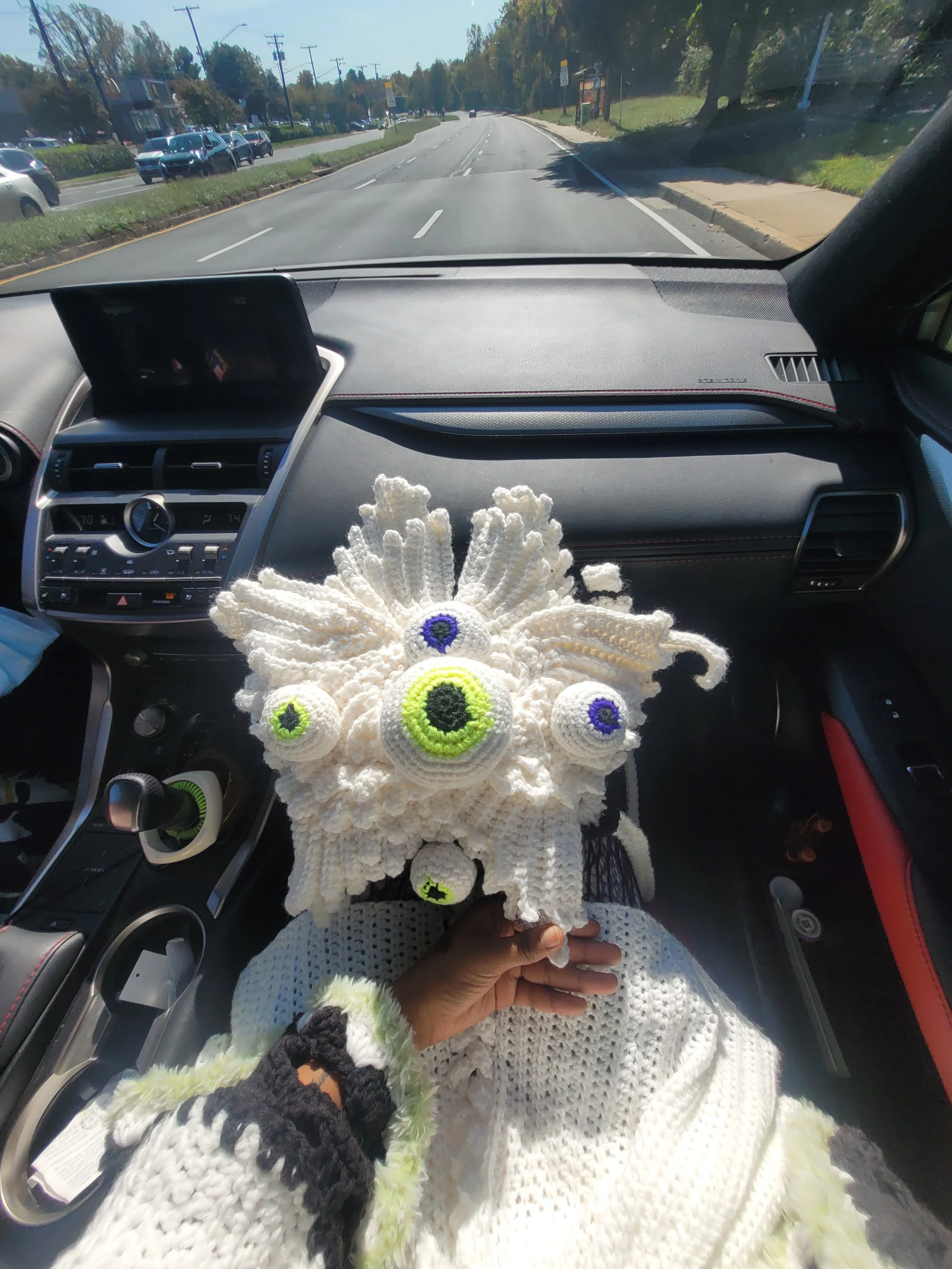 Inside a car on a sunny day, a handmade crochet creature with multiple eyes and tentacle-like appendages is being held by a person wearing a white crocheted sweater with black and neon green accents. The dashboard and part of the street are visible o