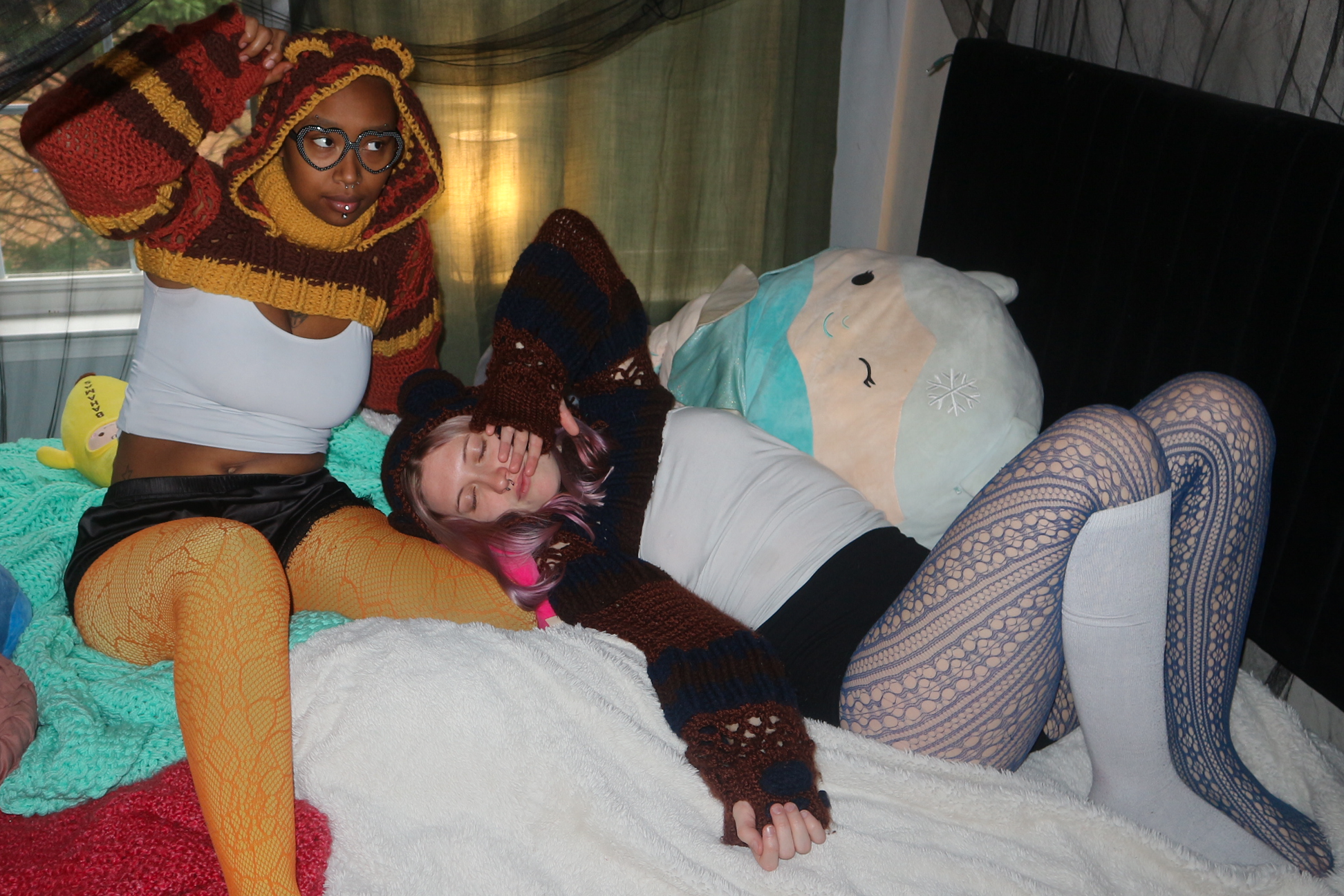 Two women in colorful, cozy clothing lying on a bed with soft blankets. One woman is sleeping, and the other is sitting beside her, looking at her with glasses and a hoodie. There is a pillow with a snowflake design and a bag of snacks on the bed.