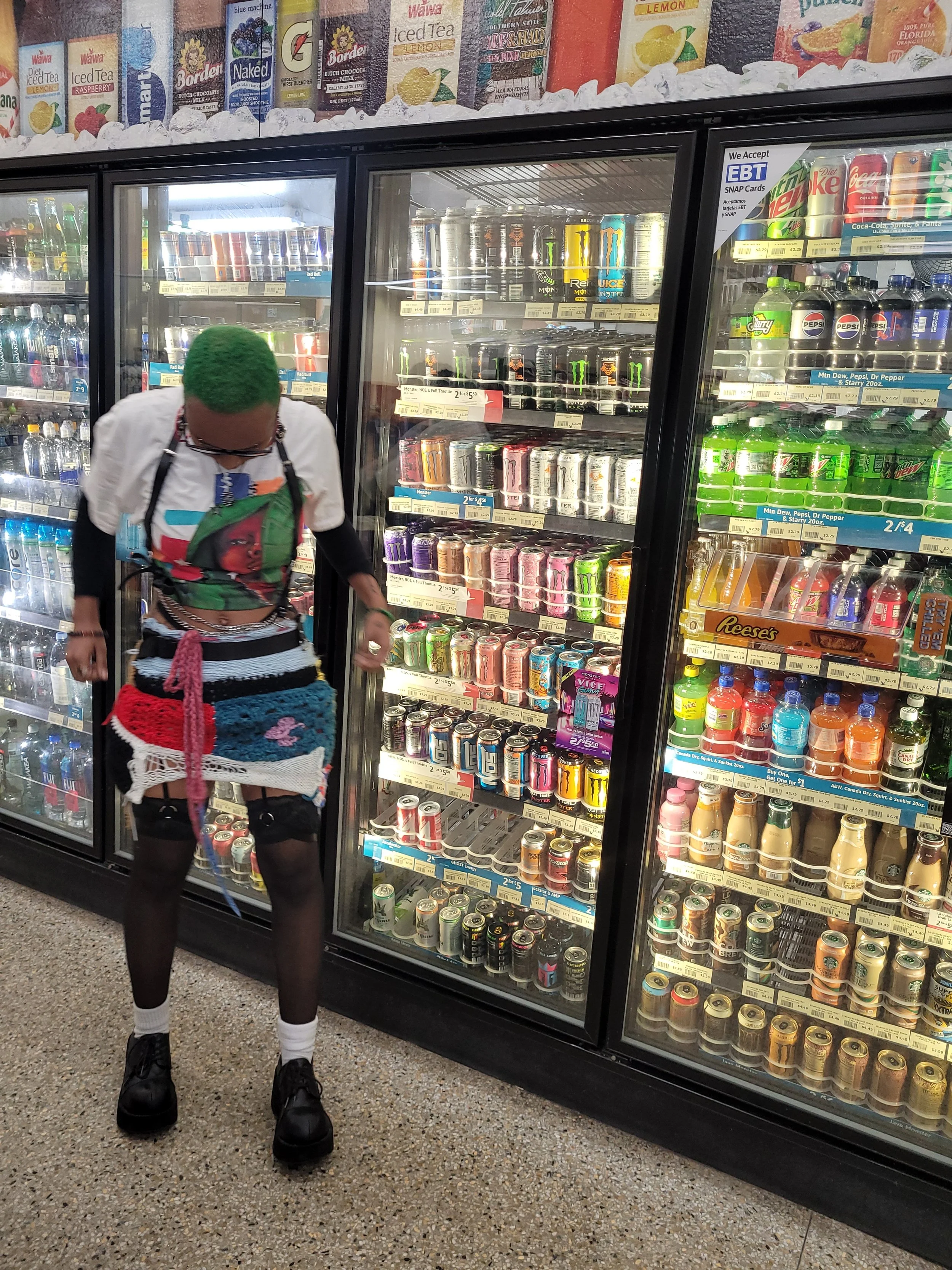 Person with bright green hair looking at soda selection in a convenience store refrigerated aisle.