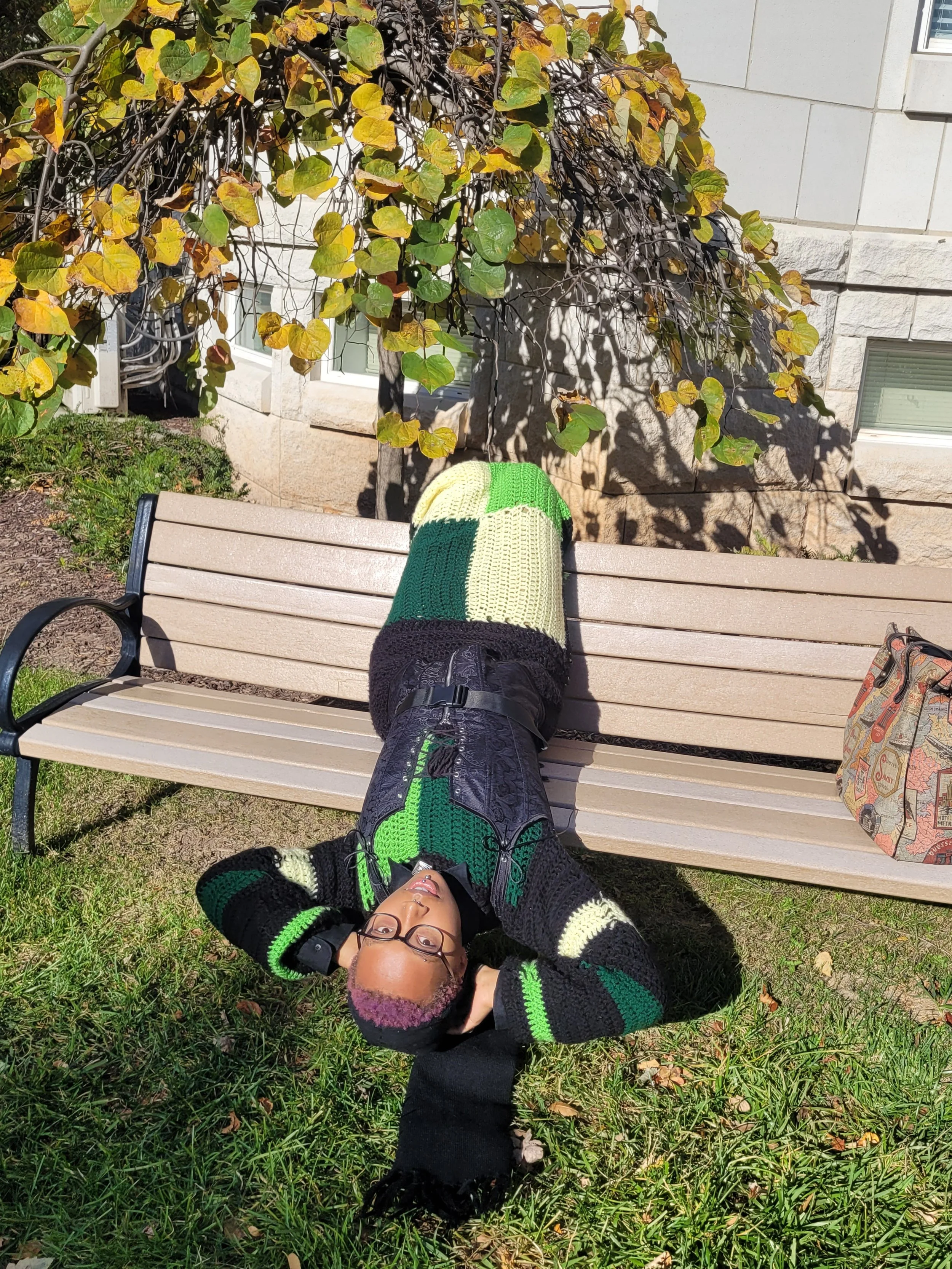 Person lying on a park bench outdoors, wearing glasses and a colorful knit sweater, resting with arms behind head, with a bag to the right and leafy plant shadowed on the building behind.