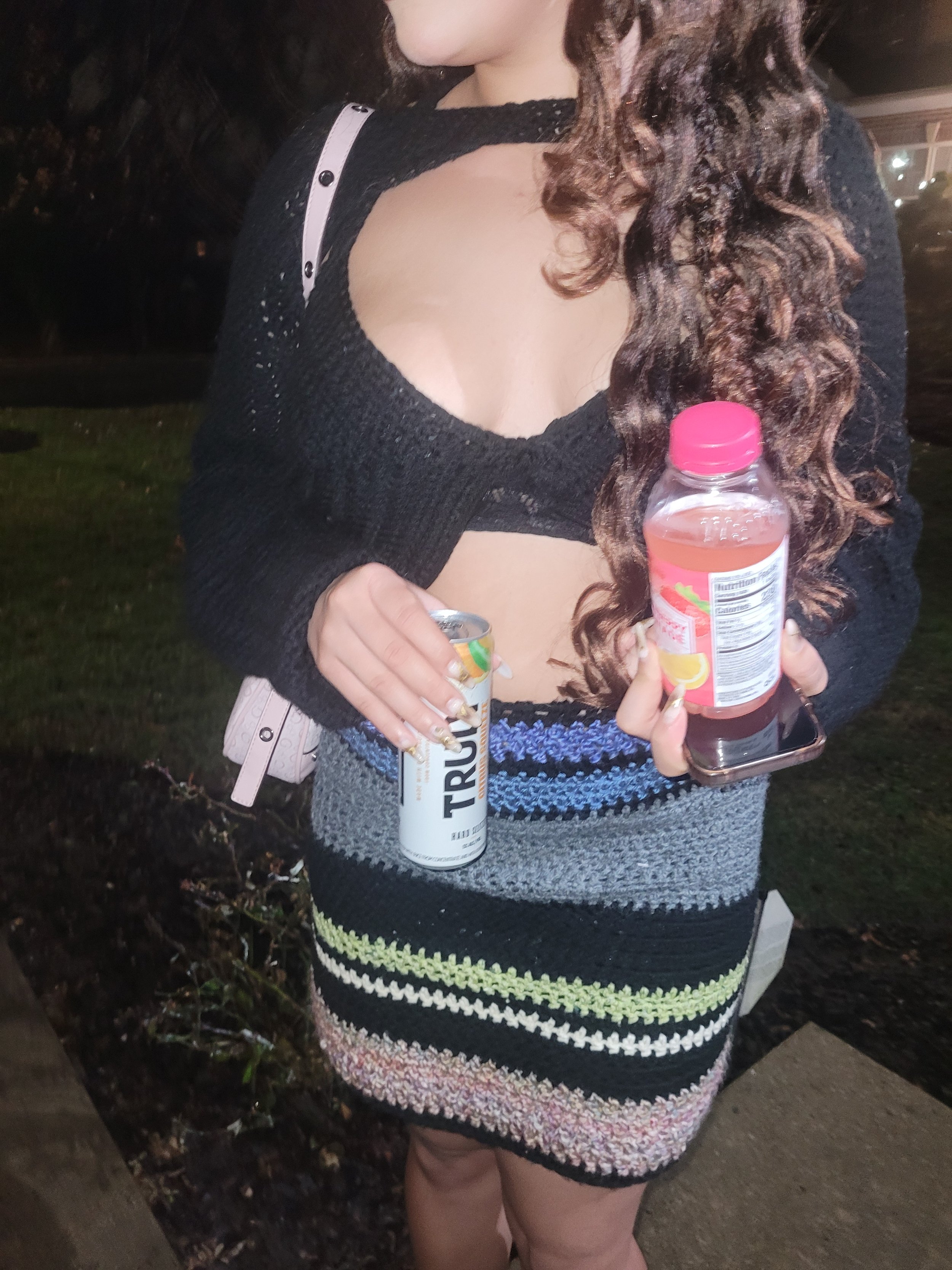A woman standing outdoors at night, holding a can of TruFruit sparkling water in her left hand and a bottle of Snapple peach iced tea in her right hand. She is wearing a black, open-knit sweater with a large cut-out on the chest and a colorful, strip