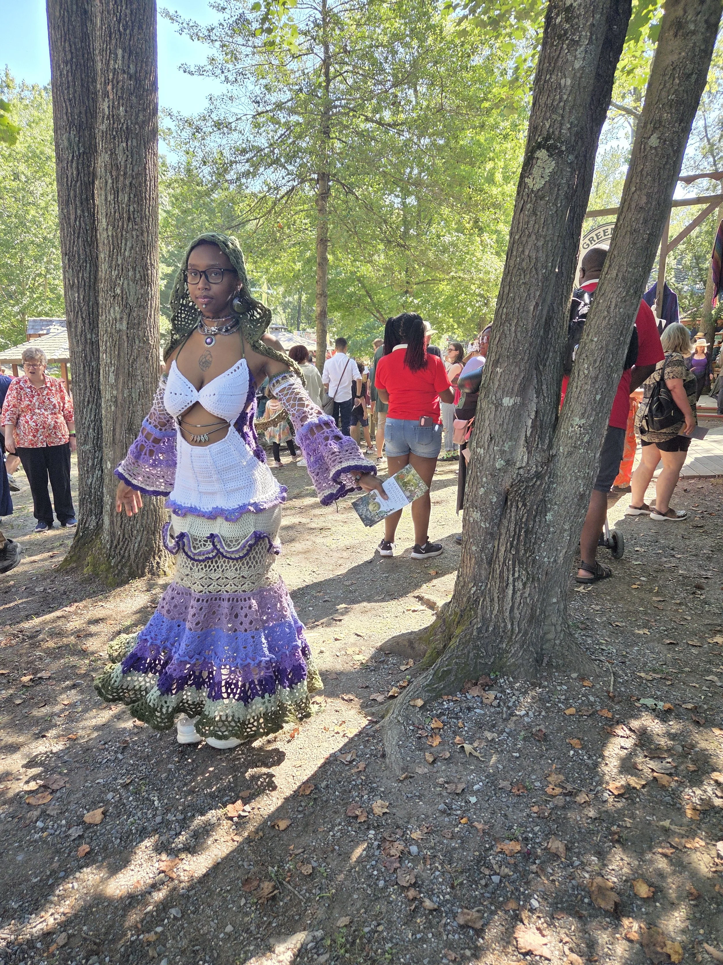 A woman wearing a colorful, crocheted, layered dress with purple, blue, green, and cream colors, standing outdoors near trees, with people and a fair or gathering in the background.