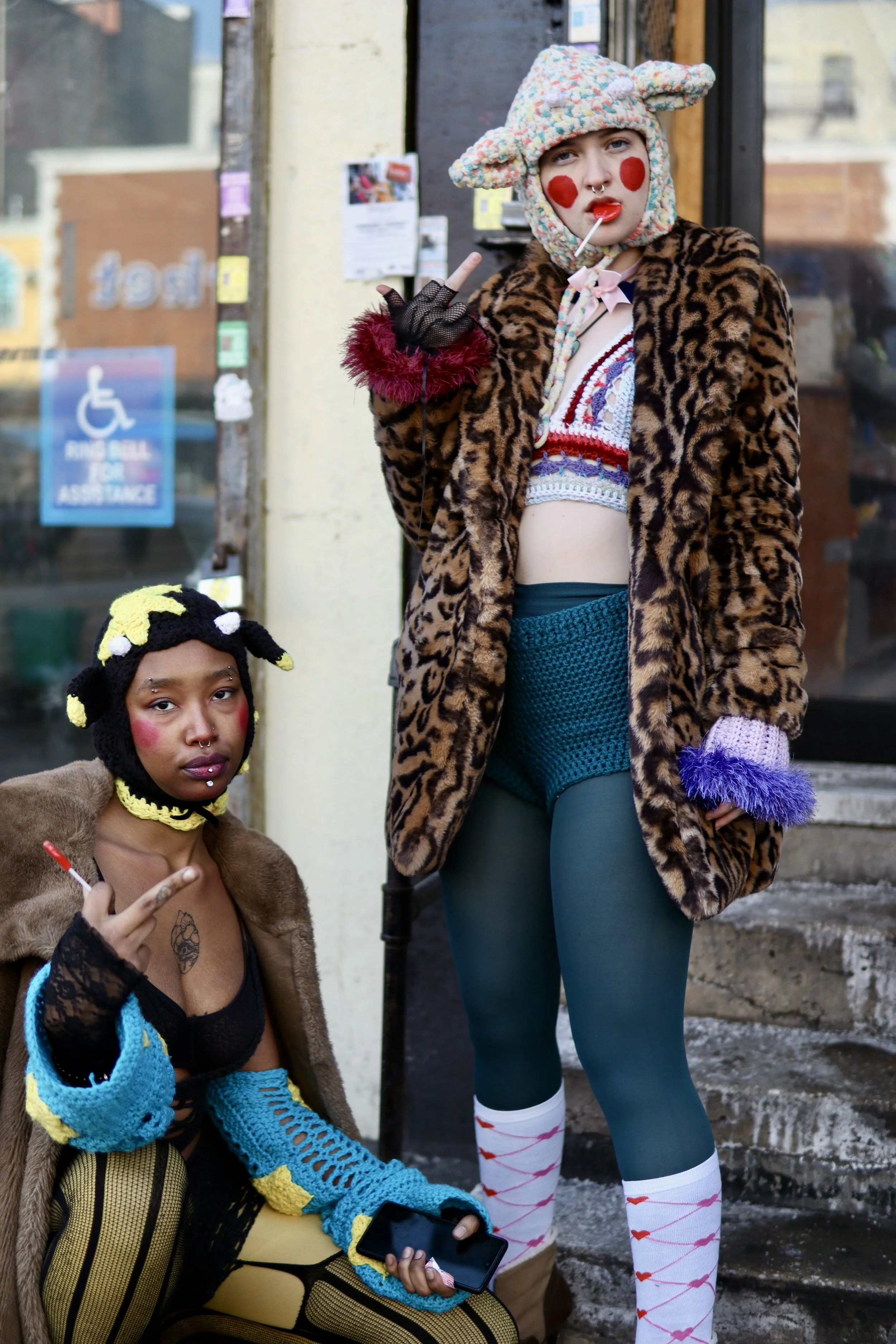 Two young women in colorful, eccentric outfits standing outside on the steps of a building. The woman standing wears a leopard print coat, a multicolored knit hat with ears, and colorful tights with a valentine's Day pattern. The woman sitting wears 