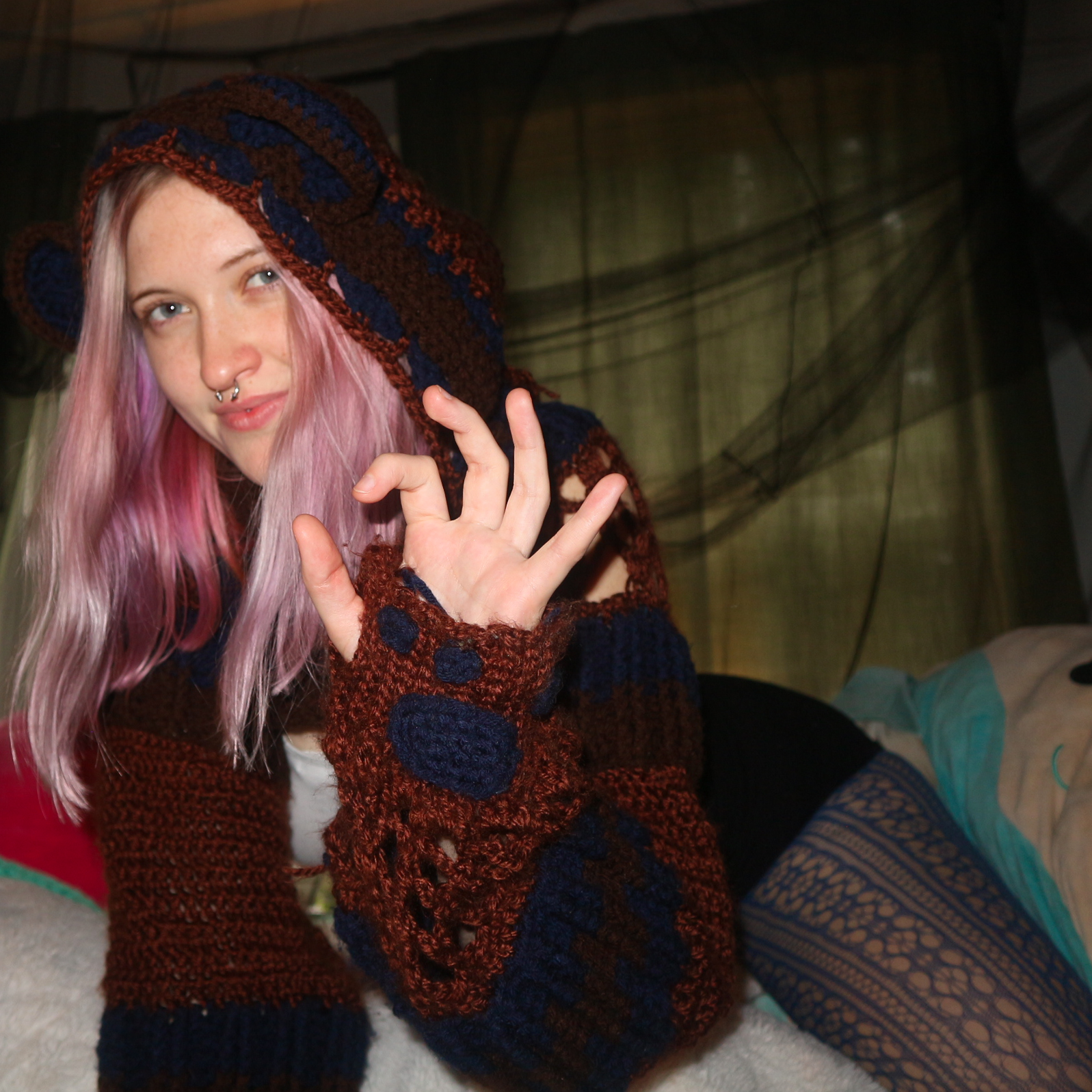 A young woman with pink hair, wearing a crocheted brown, blue, and red hooded sweater with fingerless gloves, making an OK hand gesture and smiling slightly.