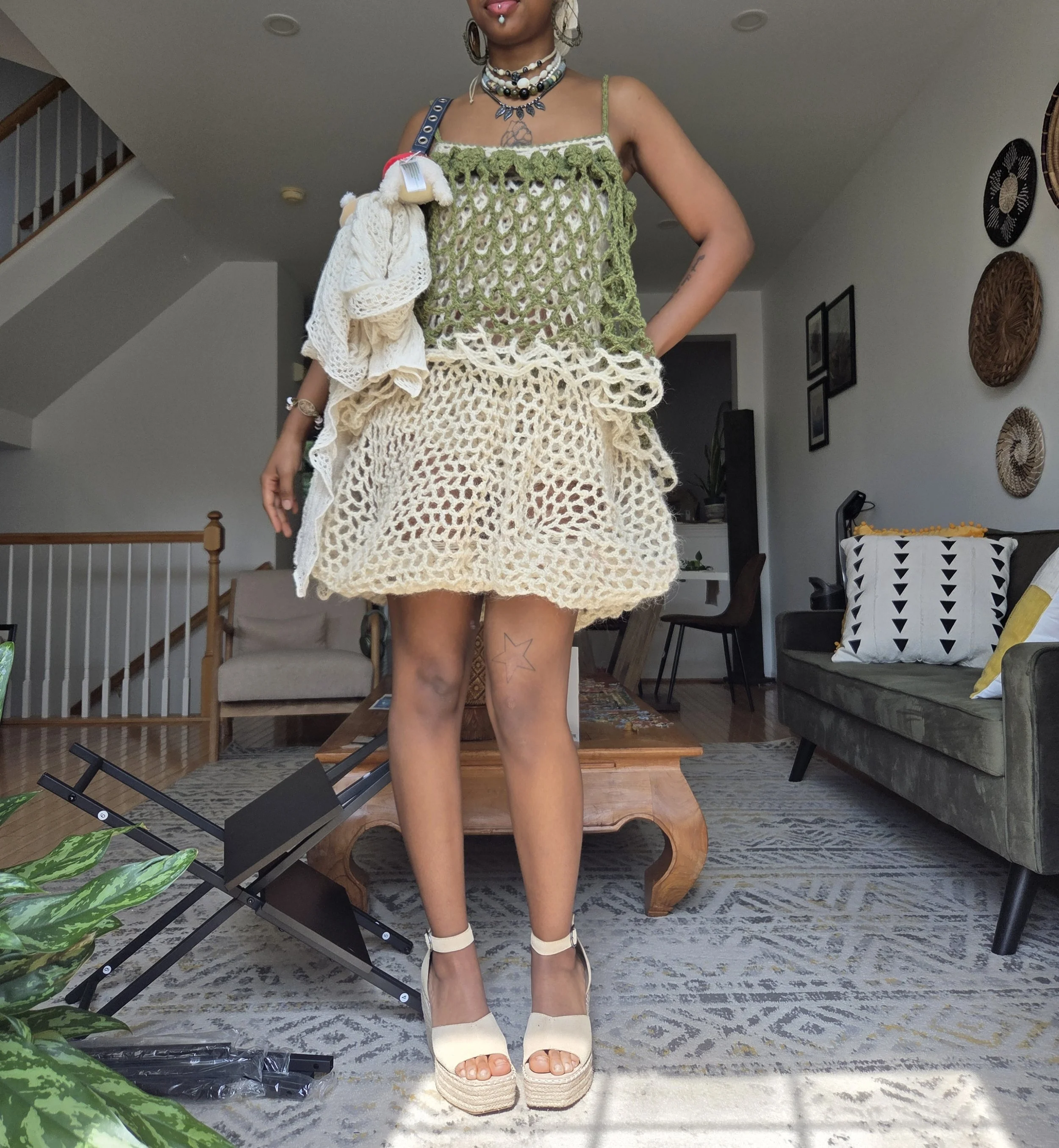 A woman standing in a living room wearing a crochet dress, platform sandals, and layered jewelry, holding a towel draped over her shoulder.