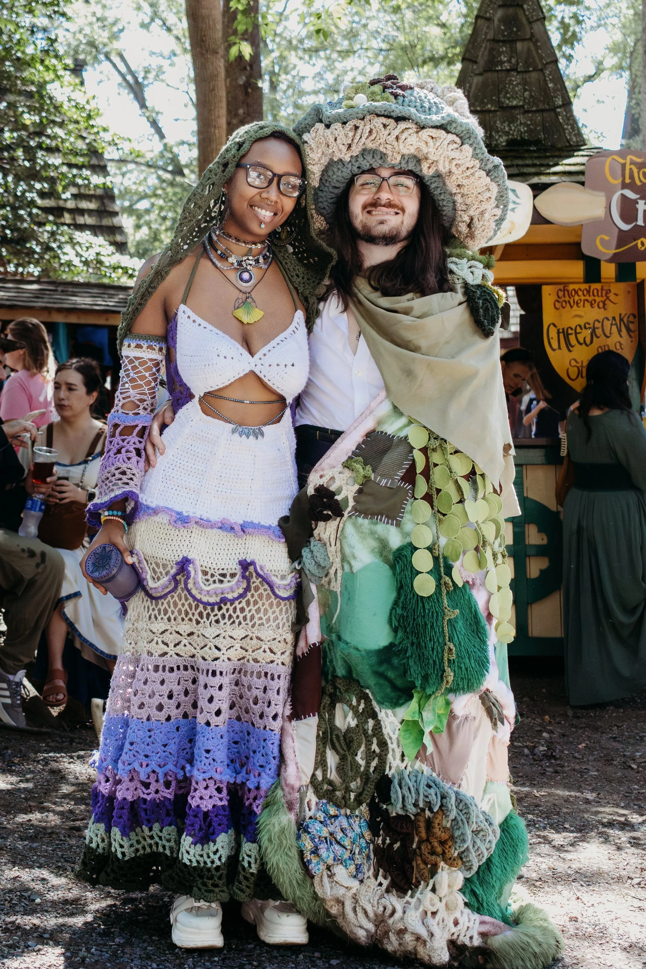 Two people standing together at an outdoor event, dressed in colorful, artistic clothing with crochet and fabric details. The woman on the left has dark skin, glasses, and is wearing a crochet dress, necklaces, and a crochet head covering. The man on