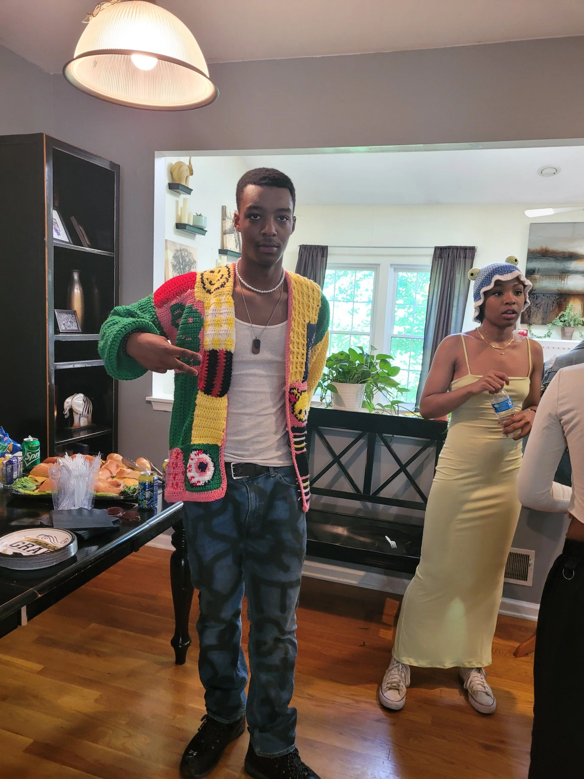 A young man with a serious expression, pointing to his colorful crochet cardigan, standing in a dining area with a table of food and two women in the background.