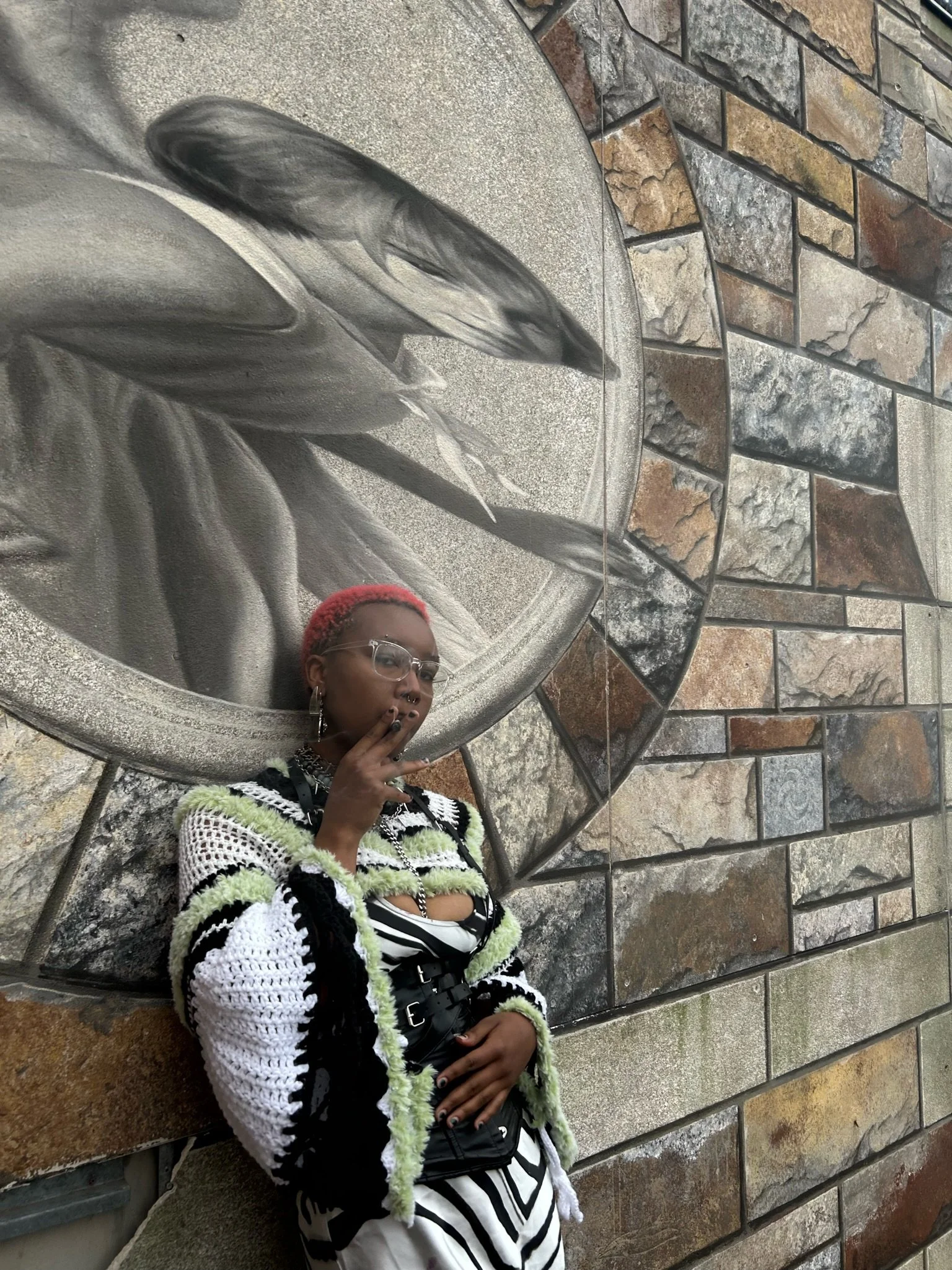 A person with short red hair and glasses stands against a stone wall with a large mural of feathers and a face. They are wearing a striped dress, a crochet jacket with green and black accents, and multiple accessories, including earrings, a necklace,