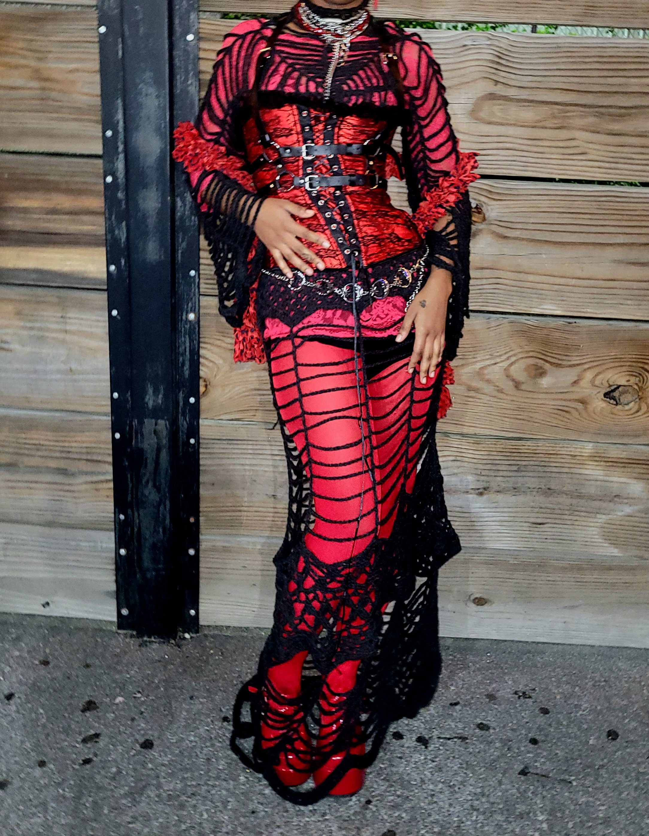 A person wearing a red and black distressed dress with black crochet overlay and red platform shoes standing against a wooden fence.