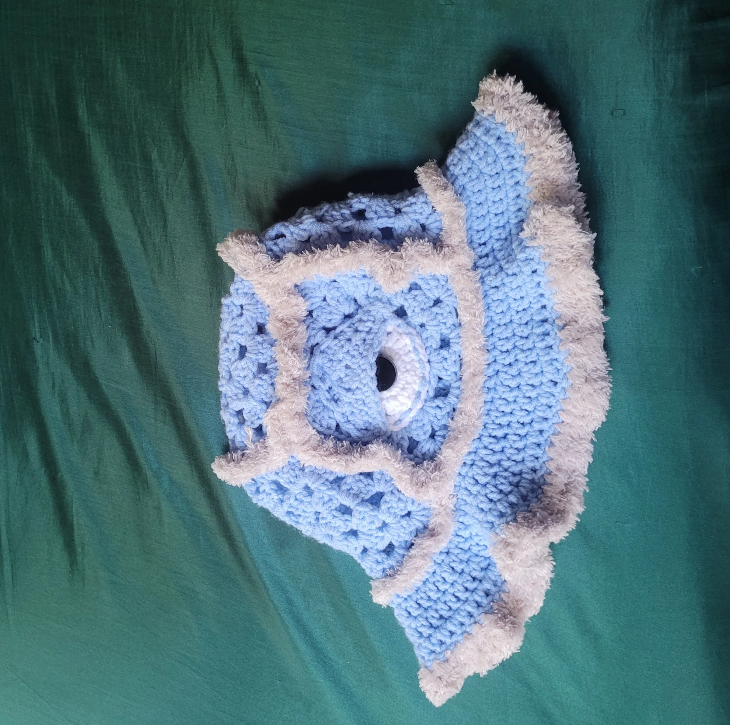 Blue crocheted sleeping mask with beige trim, resting on a green surface.