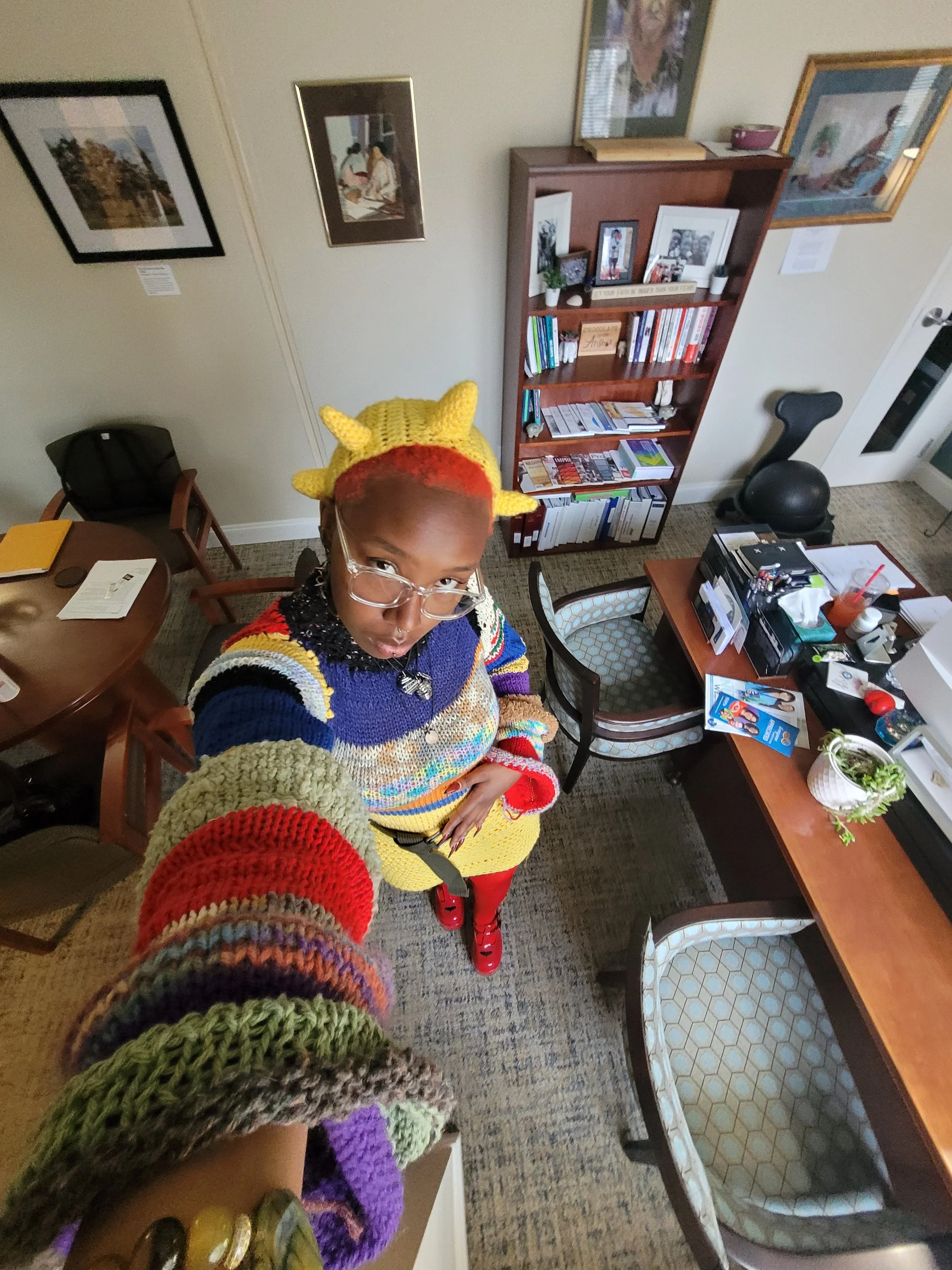 A woman with colorful hair and glasses taking a selfie in an office room decorated with framed artwork and a wooden bookshelf filled with books and photos.