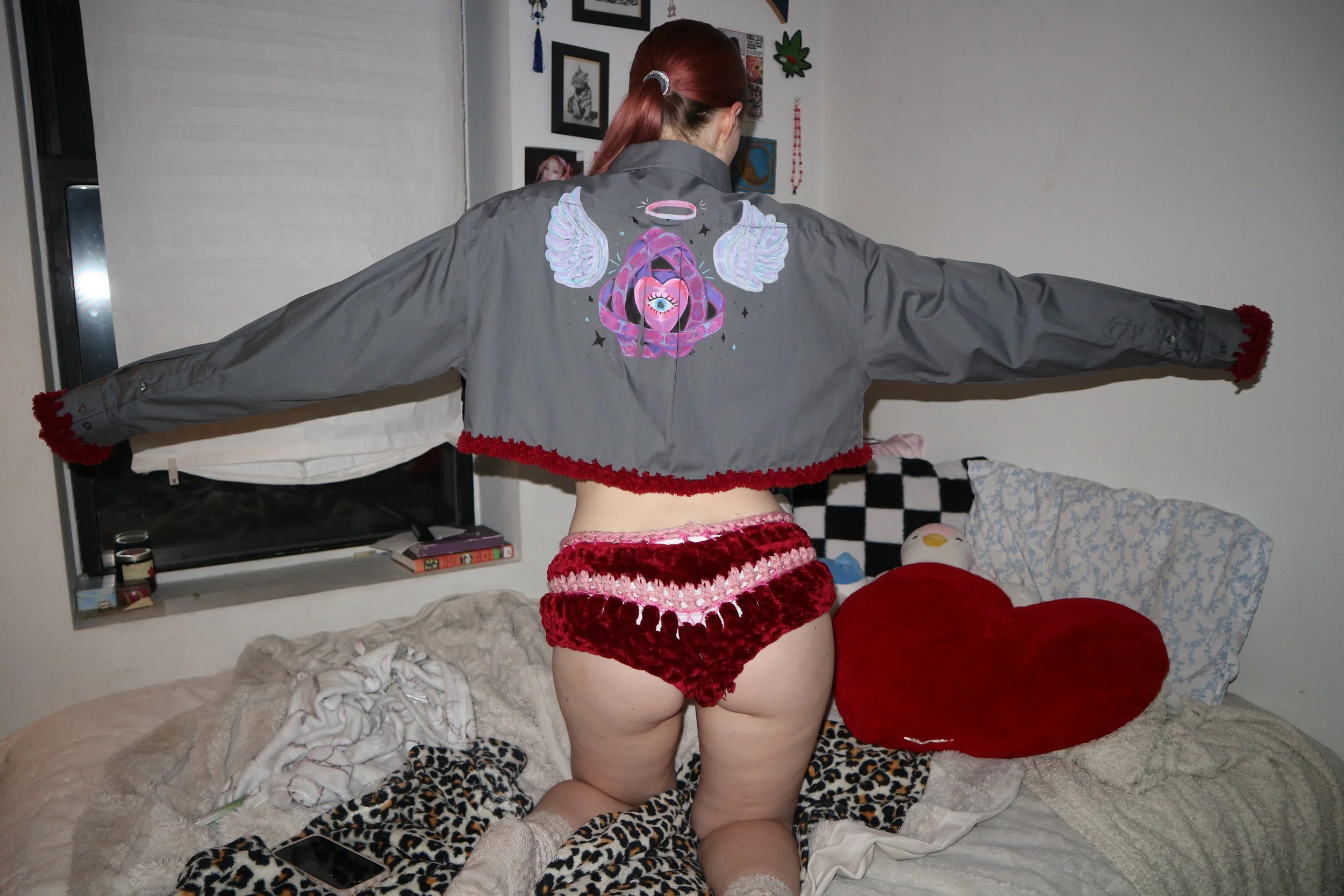 Person with red hair wearing a gray jacket with colorful graphic and angel wings, Pajama shorts, kneeling on bed with arms outstretched.