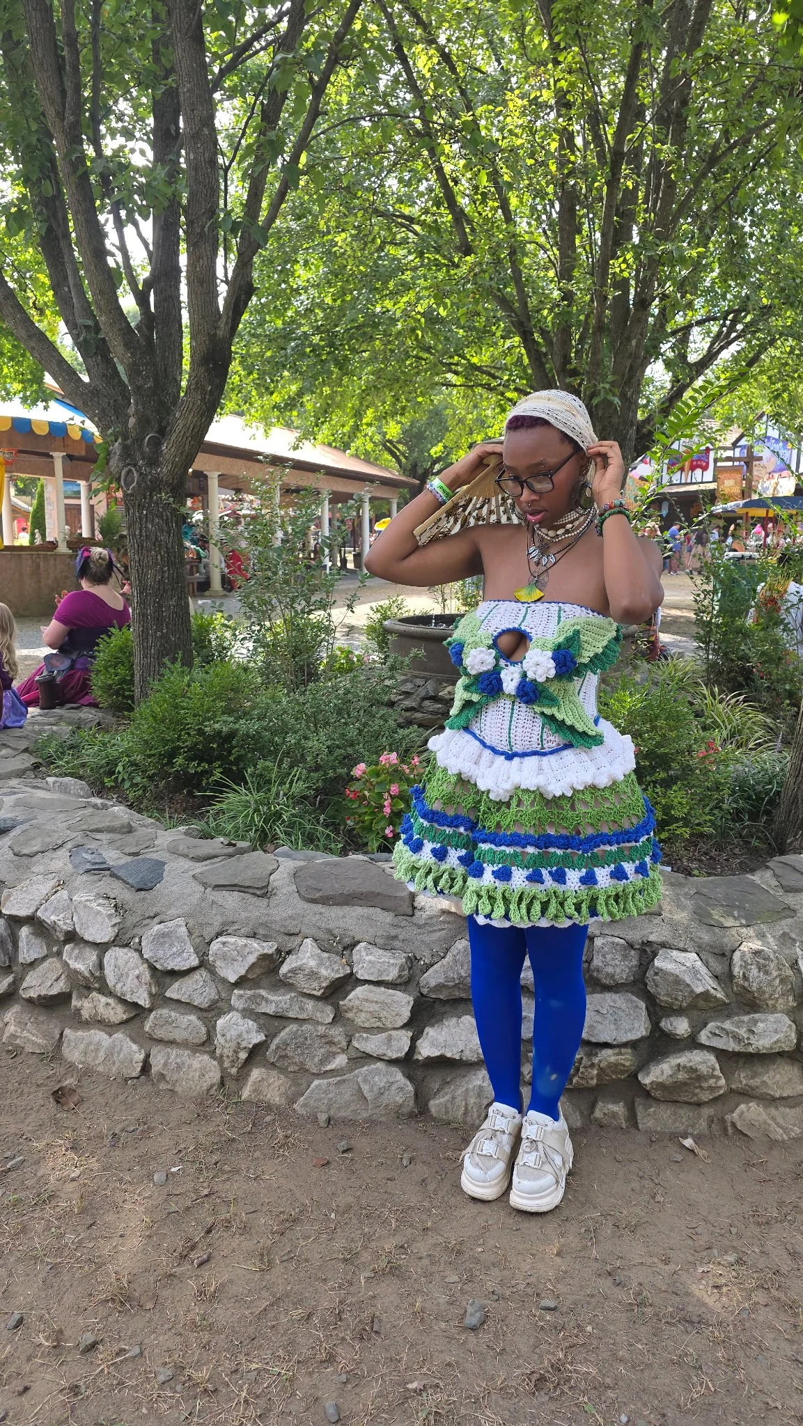 A person dressed in a colorful, handmade, crocheted outfit with blue tights and white shoes, standing outdoors near a stone wall and green trees.