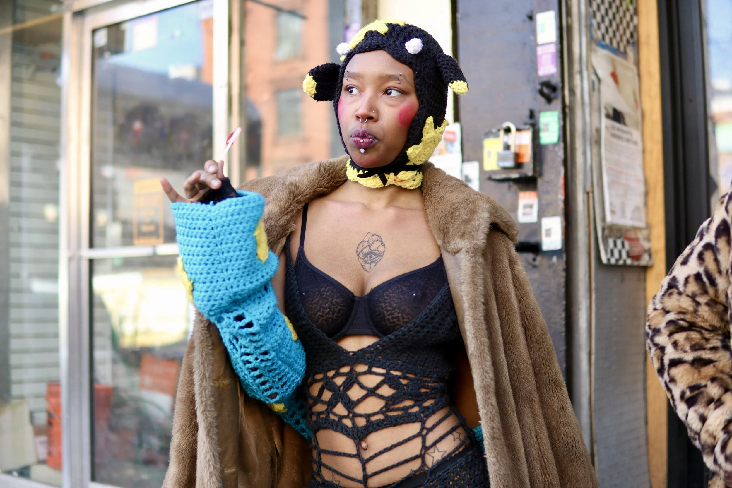 Young woman with colorful crochet glove, wearing a costume with crochet black patterns and a fur coat, has a septum piercing and a tattoo on her chest, standing on a city street.