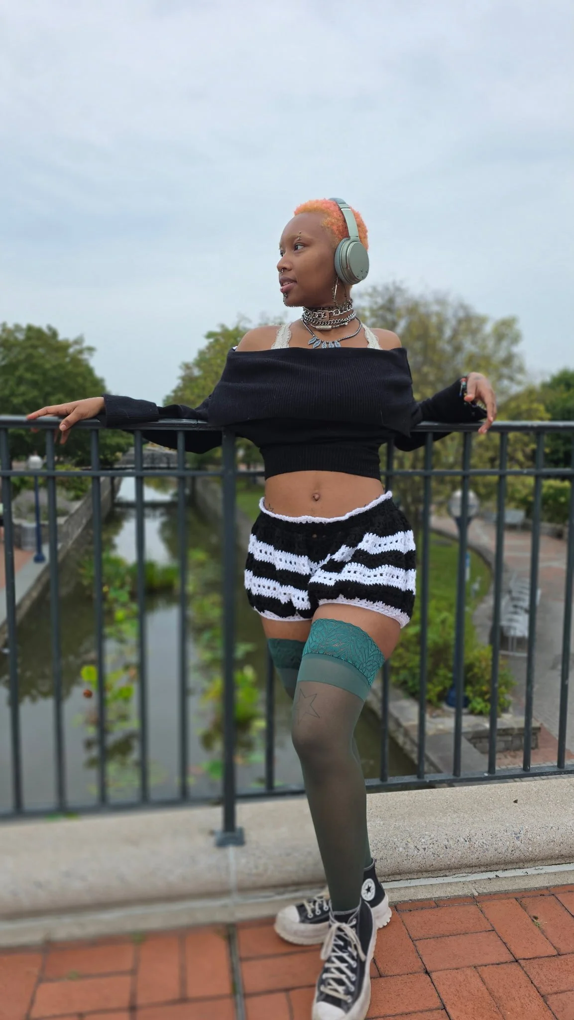 A person with short, colorful hair wearing headphones, a black off-the-shoulder sweater, black and white striped shorts, patterned thigh-high stockings, and black Converse sneakers, posing on a bridge over a river with trees in the background.