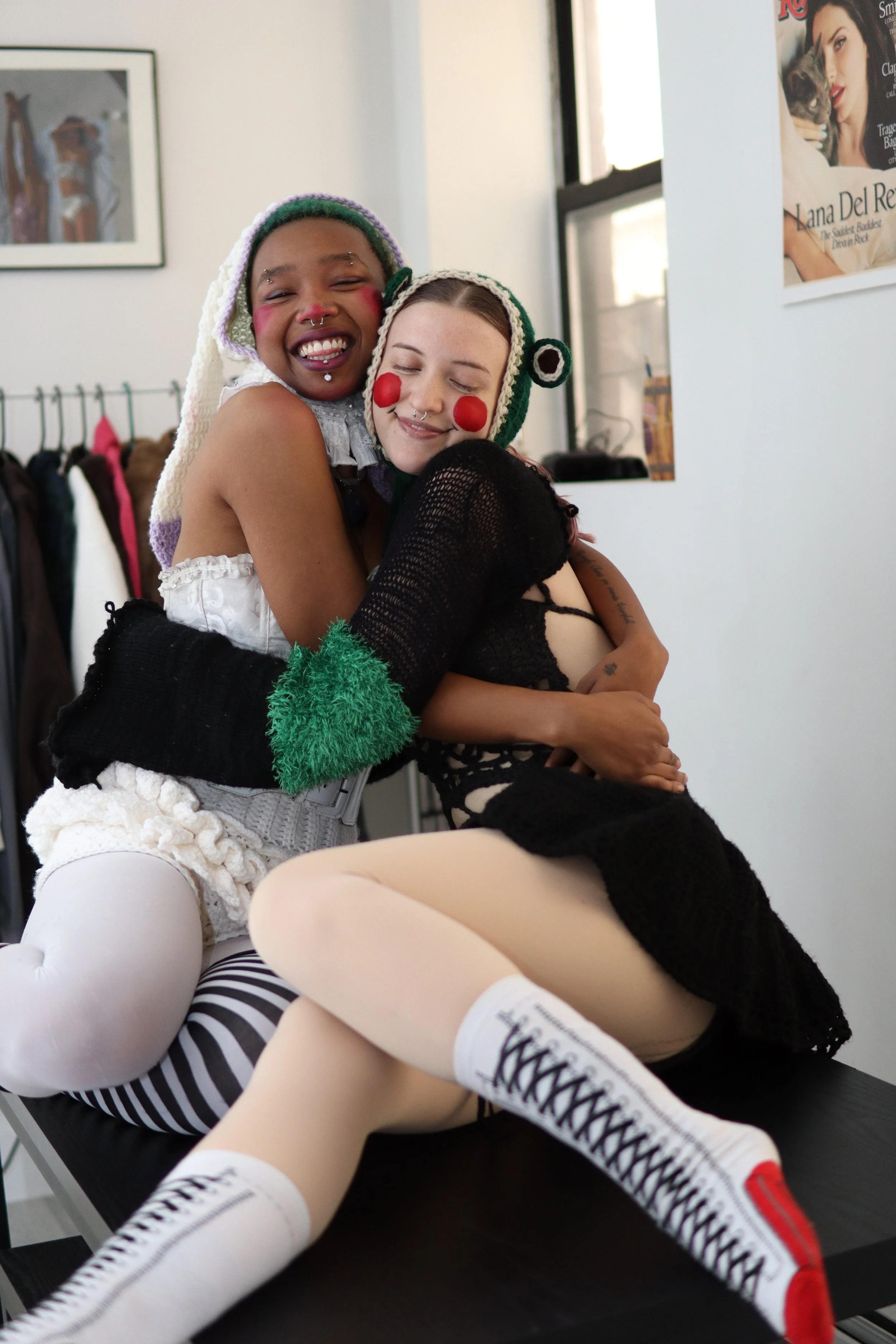 Two women dressed in playful, colorful, and revealing costumes hugging and smiling in an indoor setting with clothes rack and framed picture in the background.