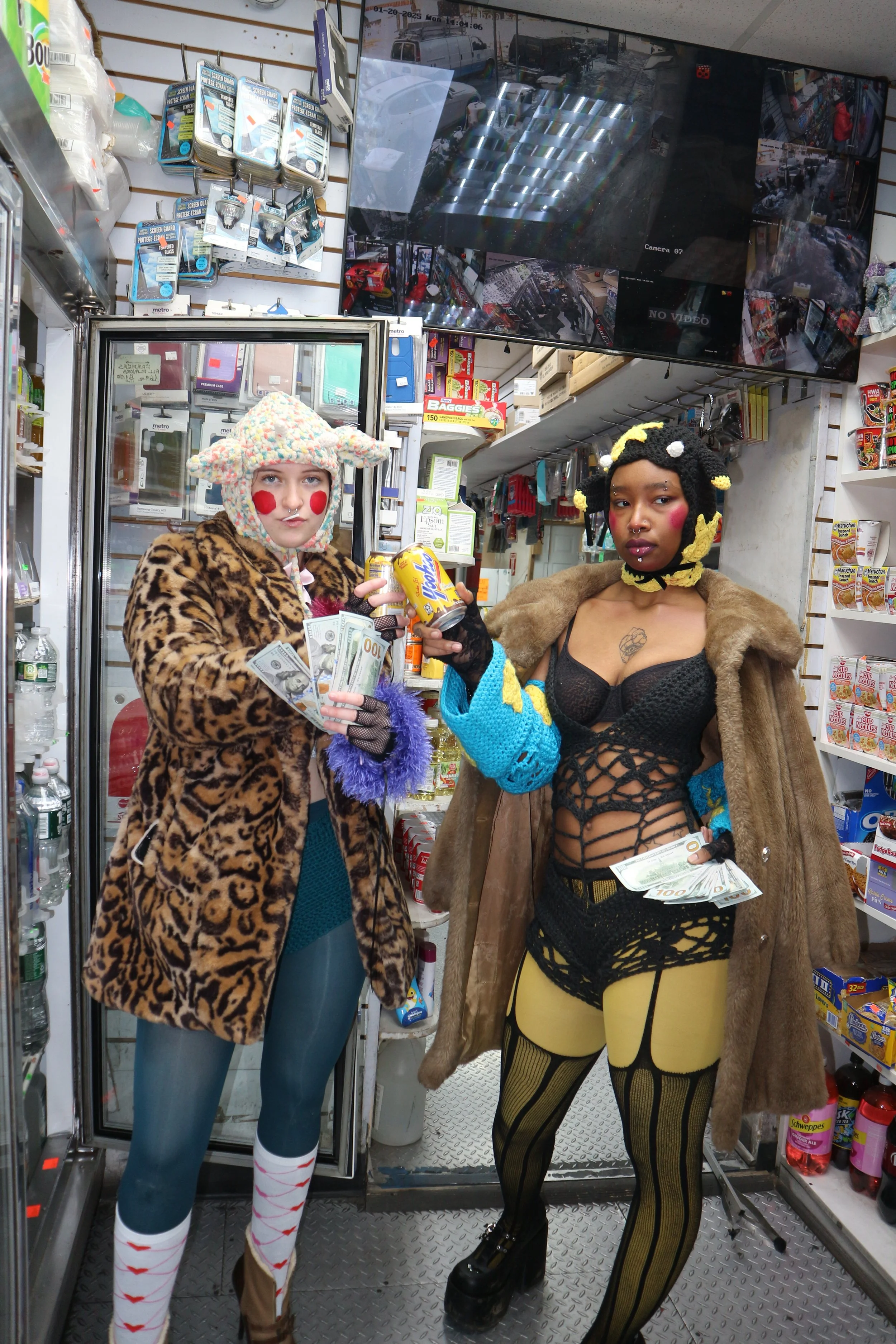 Two women in eclectic outfits standing inside a convenience store. One wears a leopard print coat and colorful knit hat, holding cash and a soda can. The other wears a fur coat over a black lace top, patterned tights, and has styled hair with yellow 