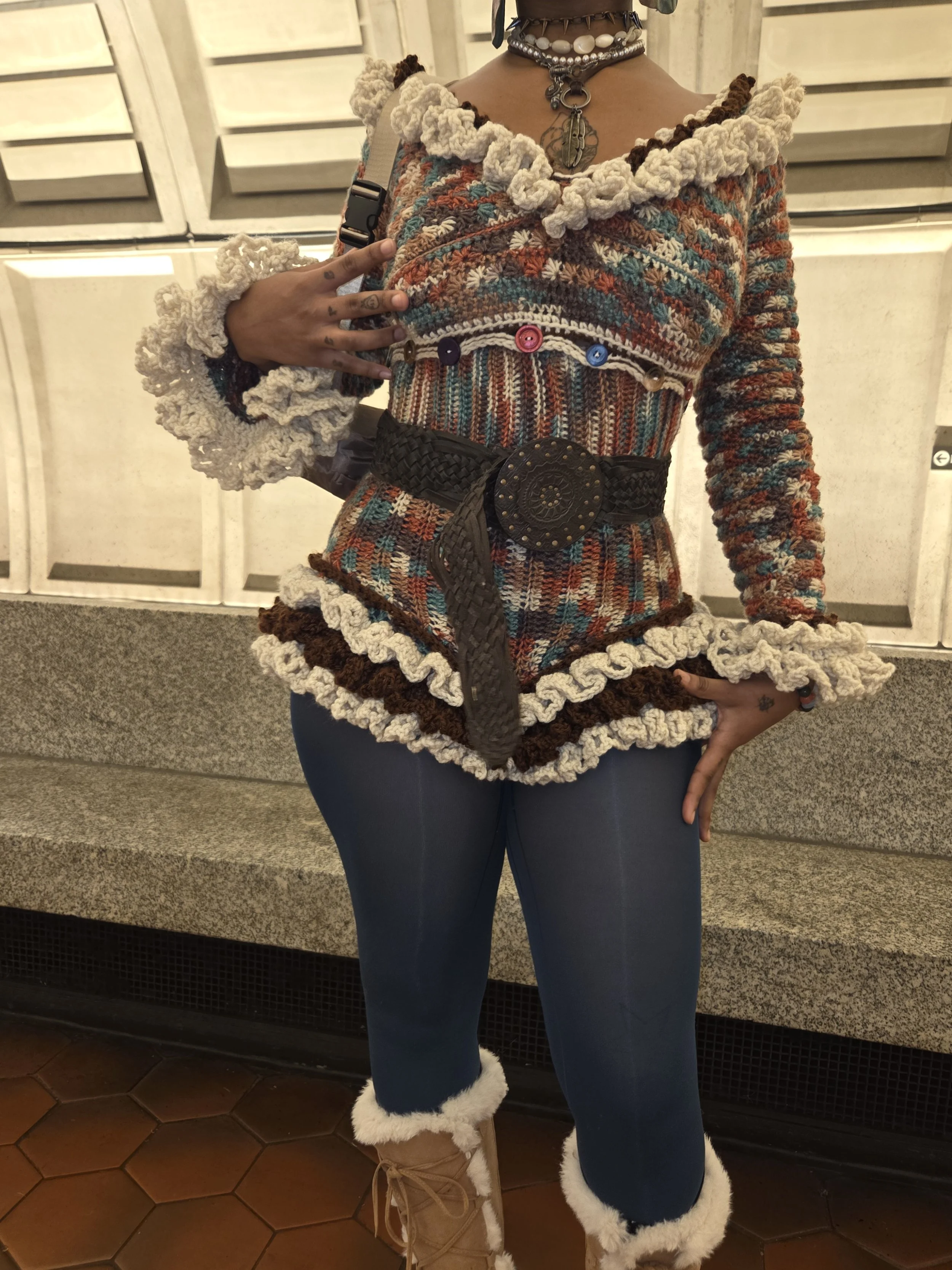 Person wearing a colorful, textured knit sweater with ruffled edges, a wide belt with a large decorative buckle, dark leggings, and tan boots with fur trim, standing against a background of metal lockers.