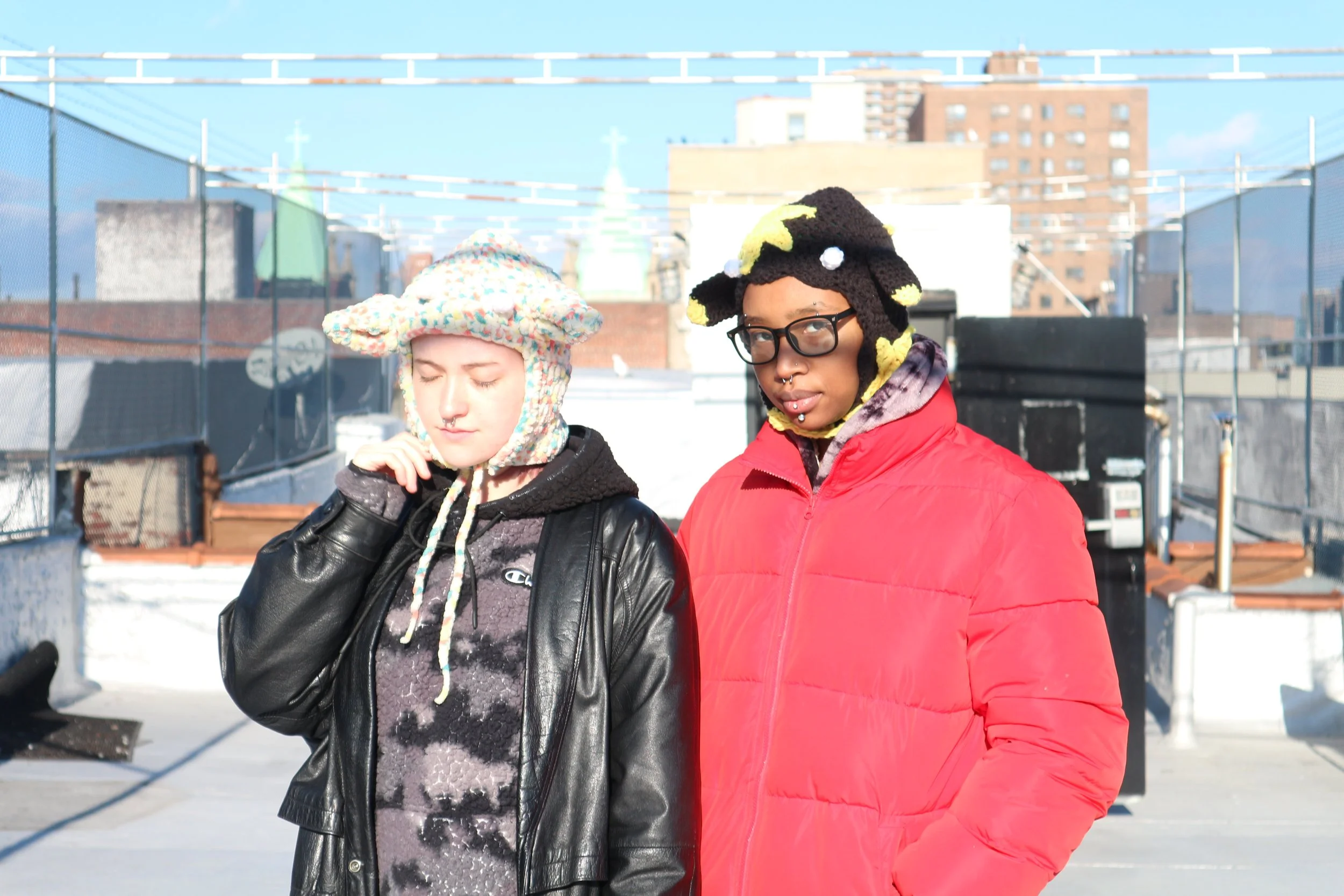 Two young women standing on a rooftop in an urban area, wearing colorful knit hats and winter jackets, with a city skyline in the background.
