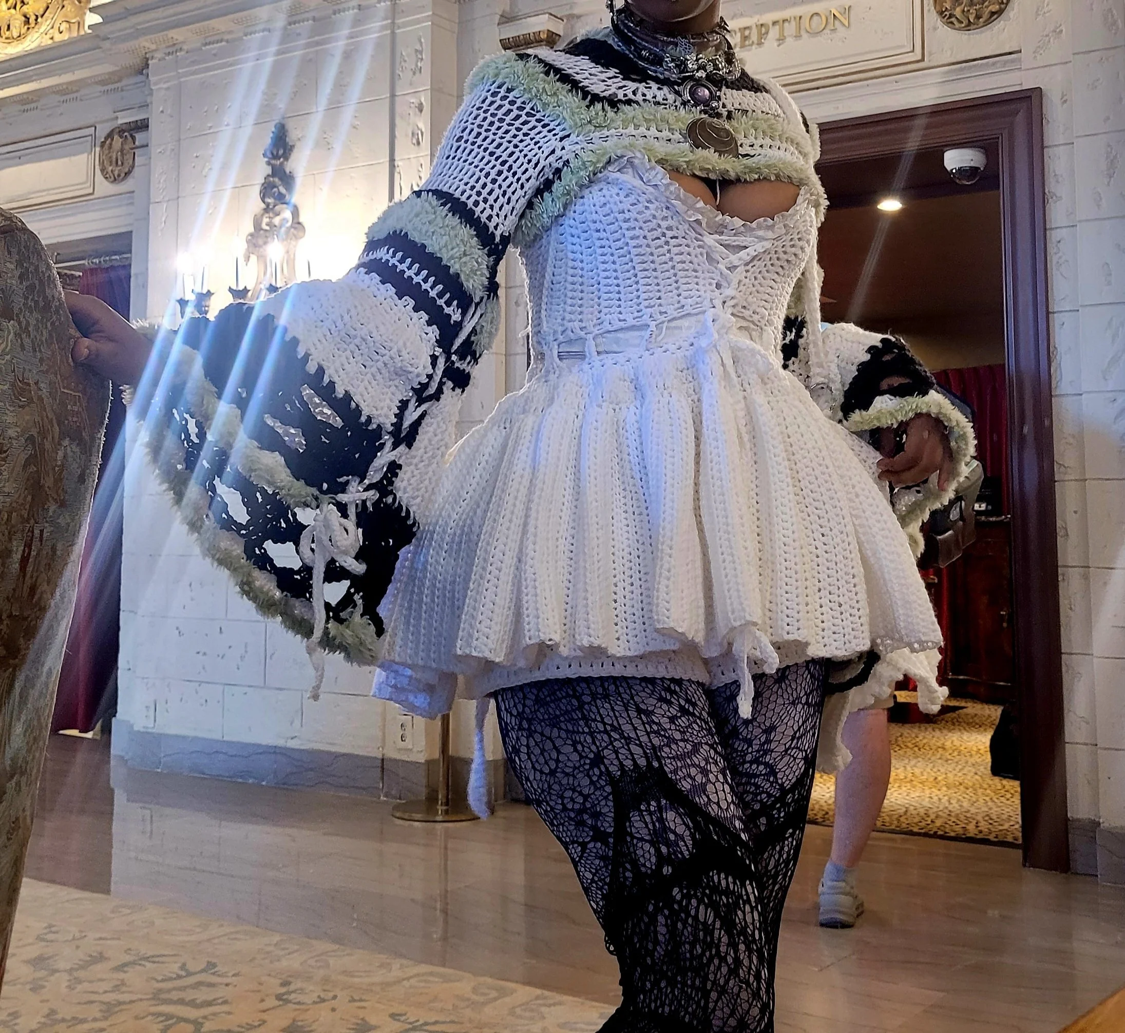 A person wearing a white, ruffled, crocheted top and black fishnet stockings with intricate web-like patterns. They are also wearing a layered crocheted shawl with stripes in black and white. The person is standing indoors in front of a reception are