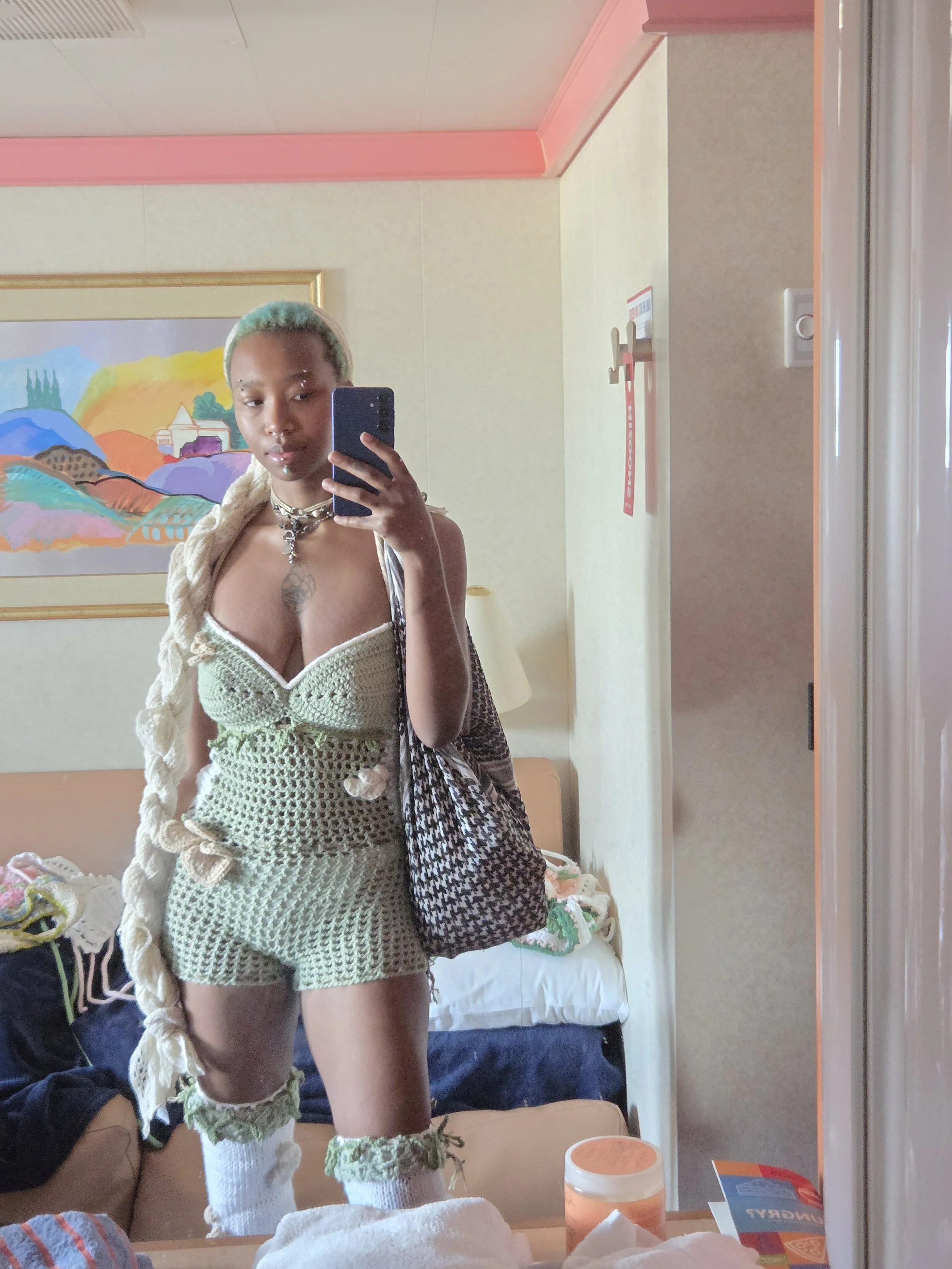 Person taking a mirror selfie in a hotel room, wearing a green crocheted outfit with knee-high socks and carrying a patterned bag, with a colorful landscape painting on the wall behind.