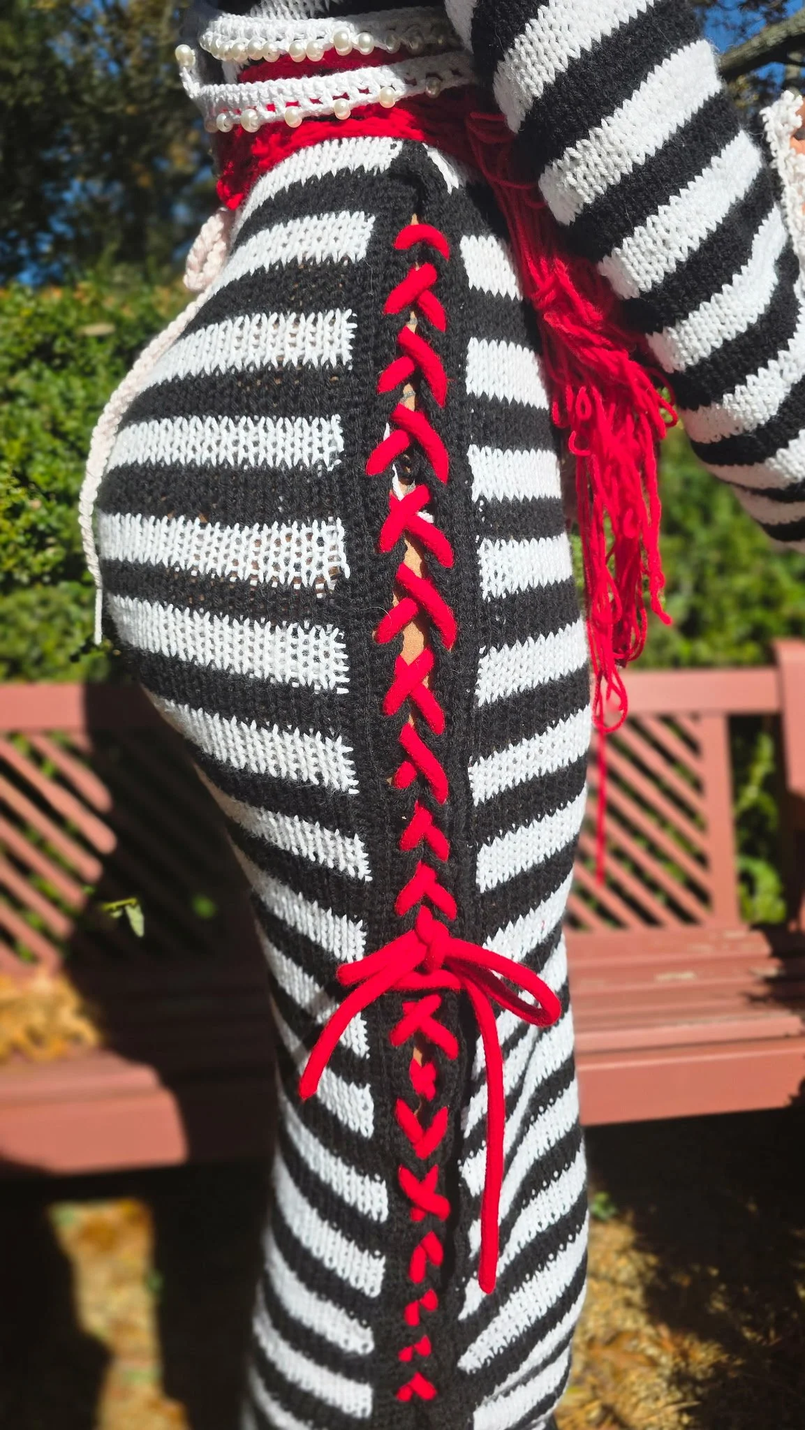 Close-up of a person's leg wearing black and white striped pants with red laces running down the side, tied at the bottom with a bow. The person is outdoors, with a pink garden bench and green foliage in the background.