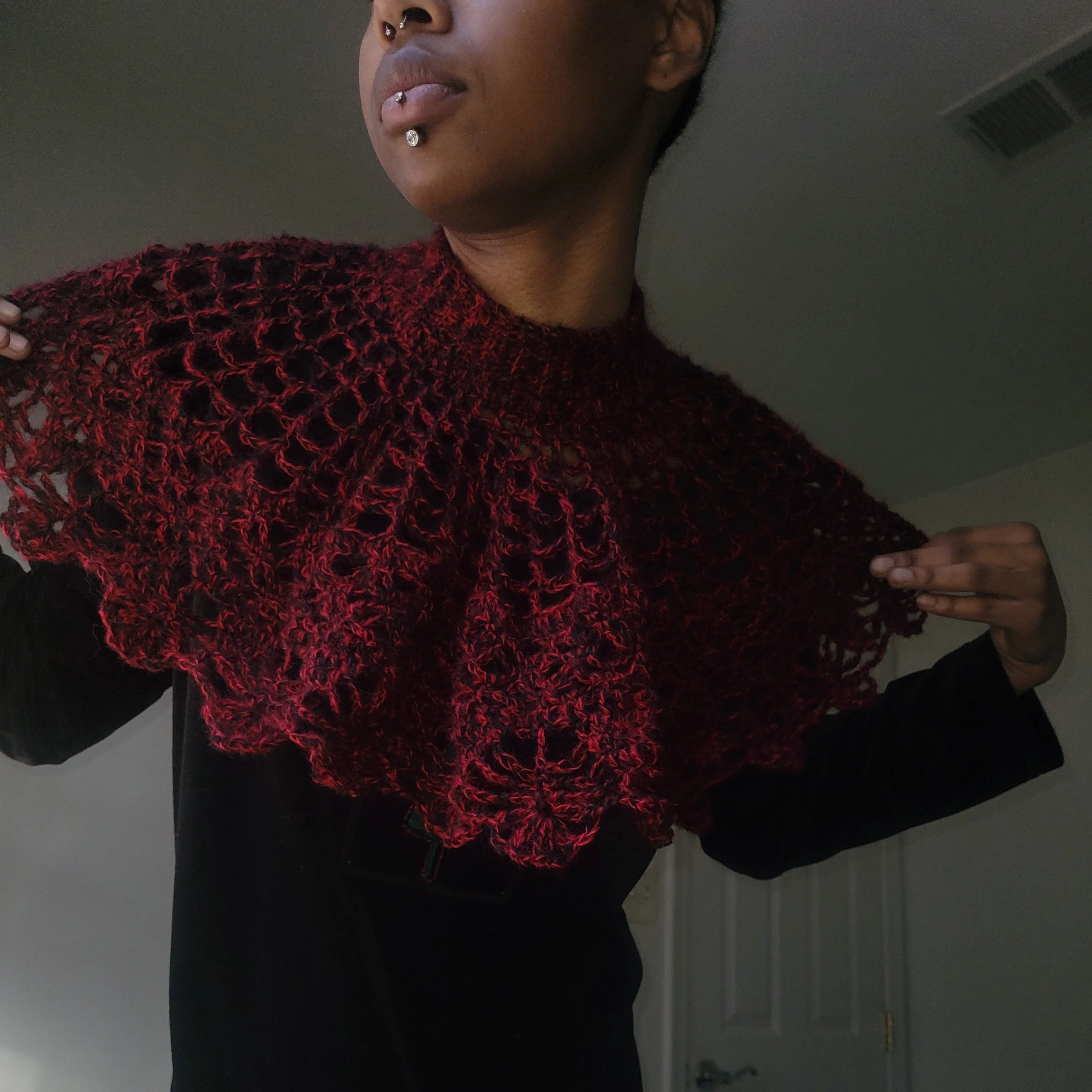 Person wearing a dark outfit with a red crocheted shawl, holding it out with both hands. The person's face is partially visible, with short dark hair and piercings on their lips.