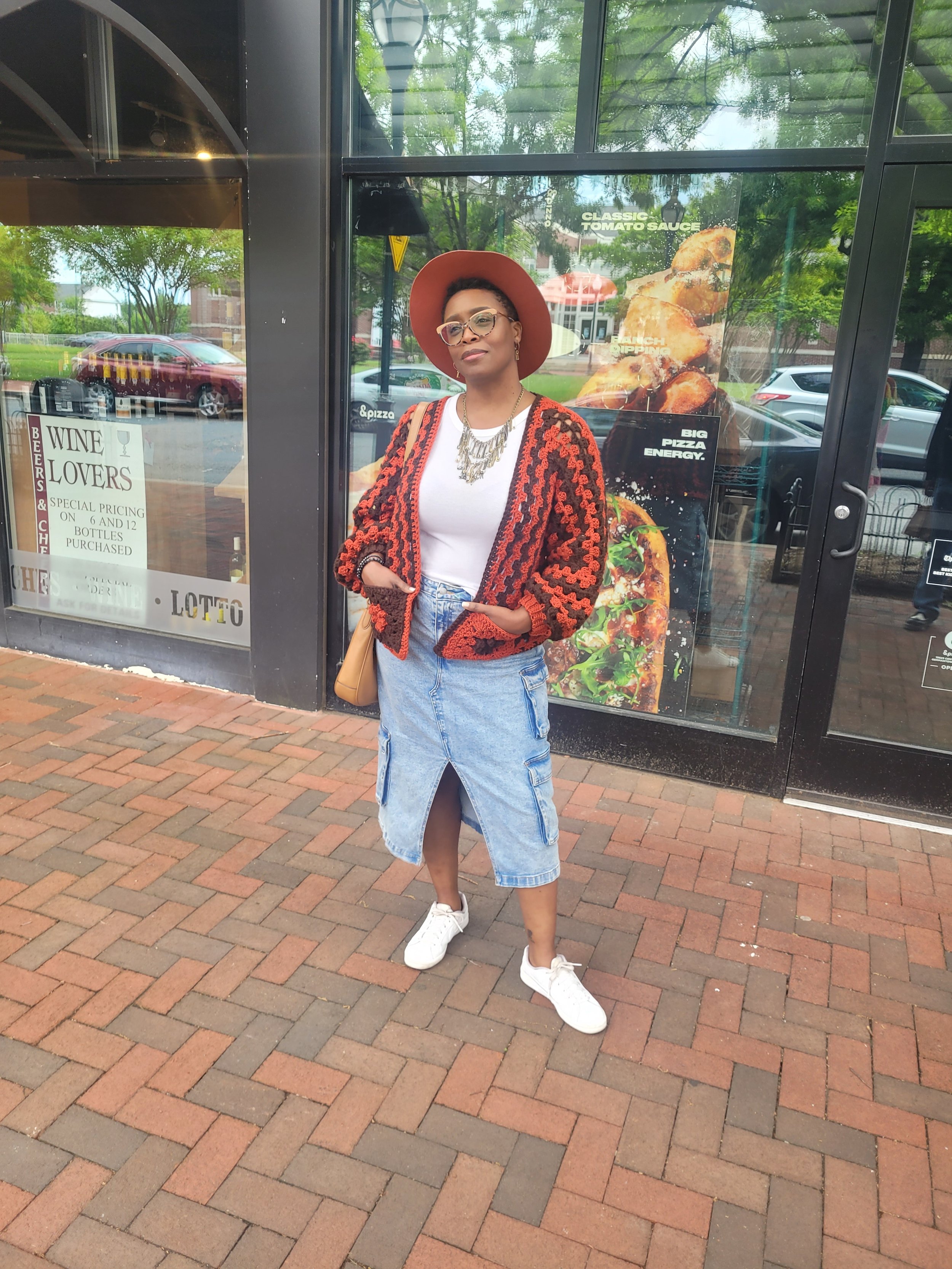 A woman standing on a brick sidewalk outside a restaurant, wearing a red hat, glasses, a gray and orange crocheted sweater, a white T-shirt, a denim skirt with a front slit, white sneakers, and carrying a tan purse.