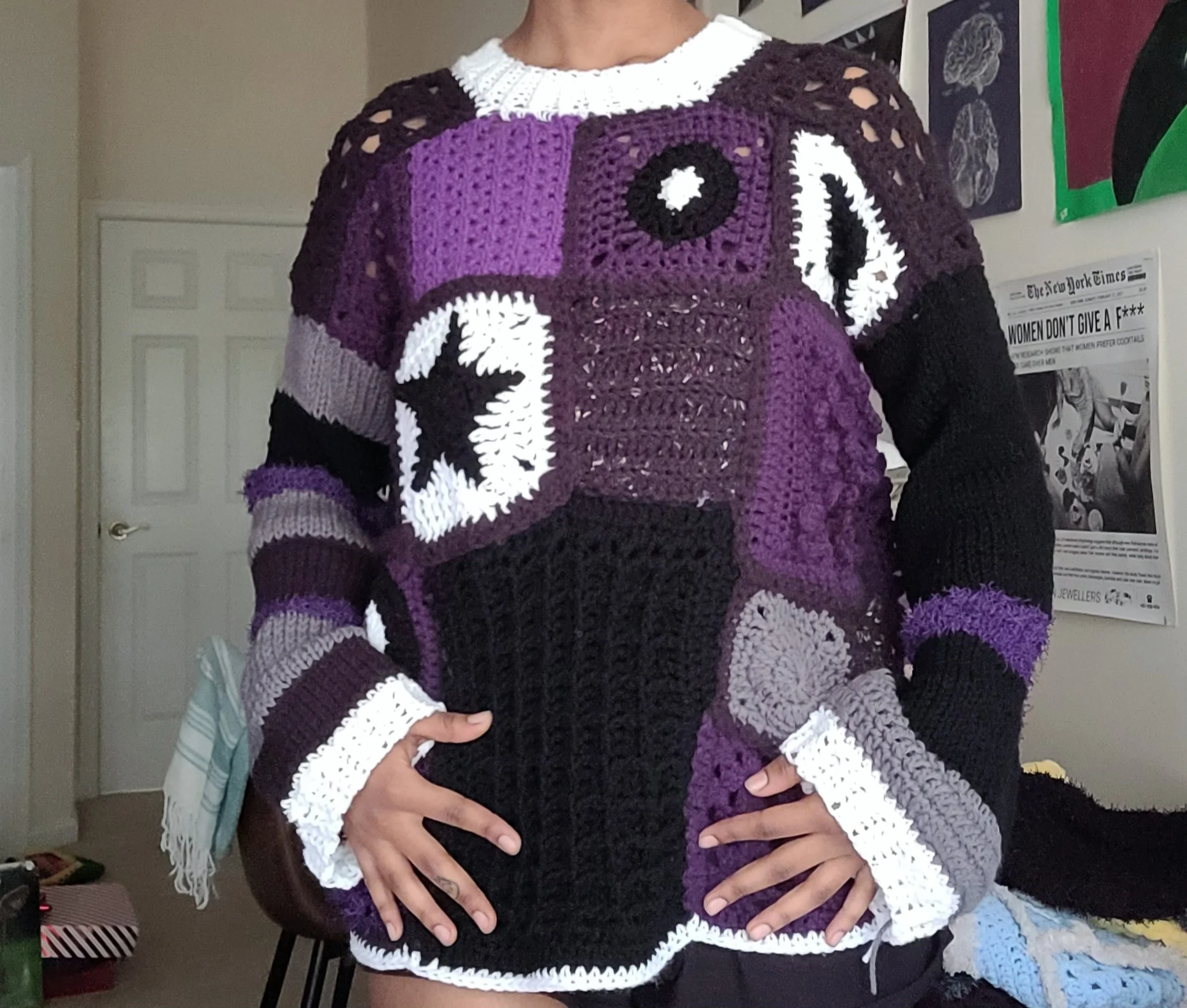 Person wearing a multicolored crocheted sweater with purple, black, white, and gray blocks and patterns, standing indoors near a wall with posters and artwork.