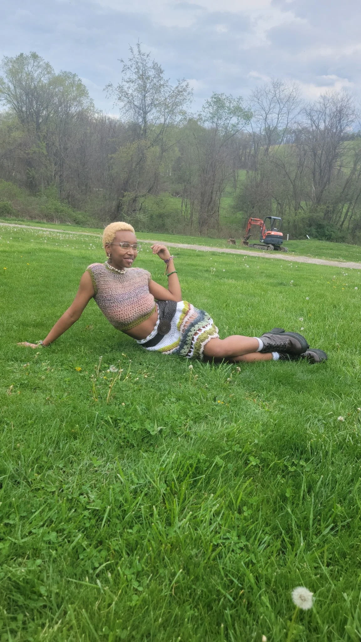 A person with short blonde hair, glasses, and wearing a multicolored crochet sleeveless top and a matching skirt, lies on grass in a field with a smile, with an excavator and trees in the background.