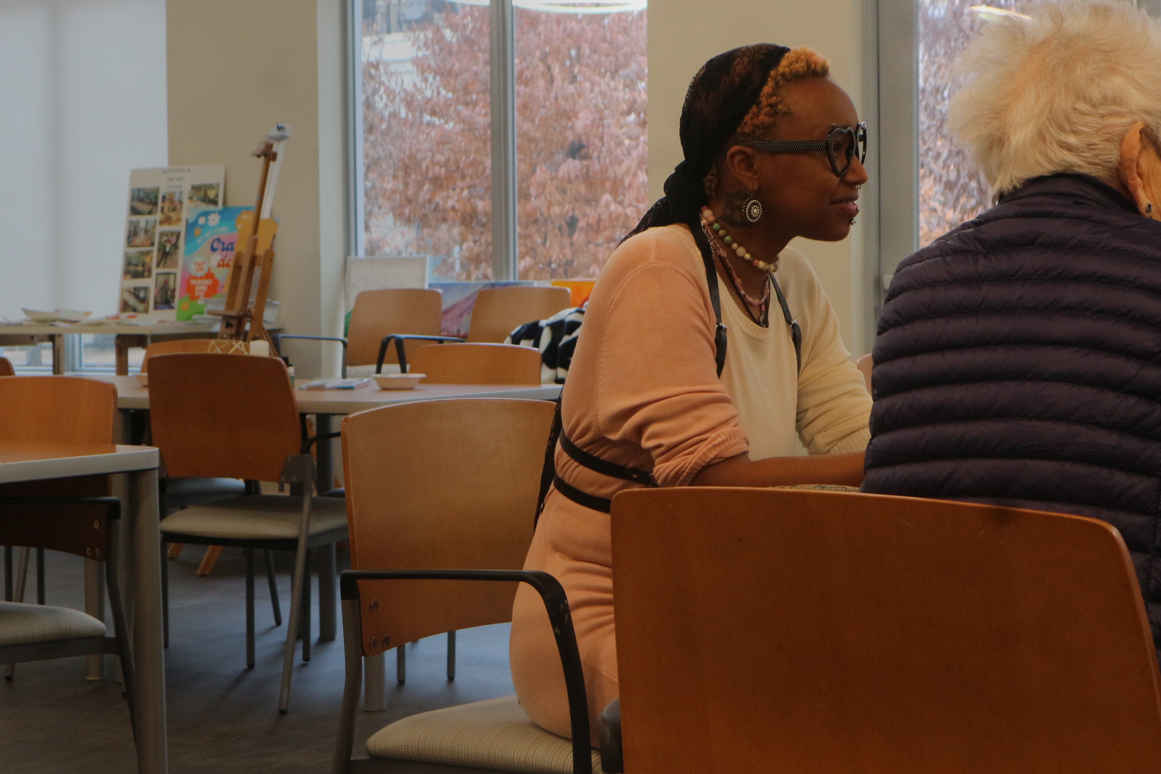 Two women sitting at a table in a brightly lit room with large windows and pink trees outside. One woman is wearing glasses, a beige shirt, and has jewelry, and the other has white hair and a dark puffer jacket.