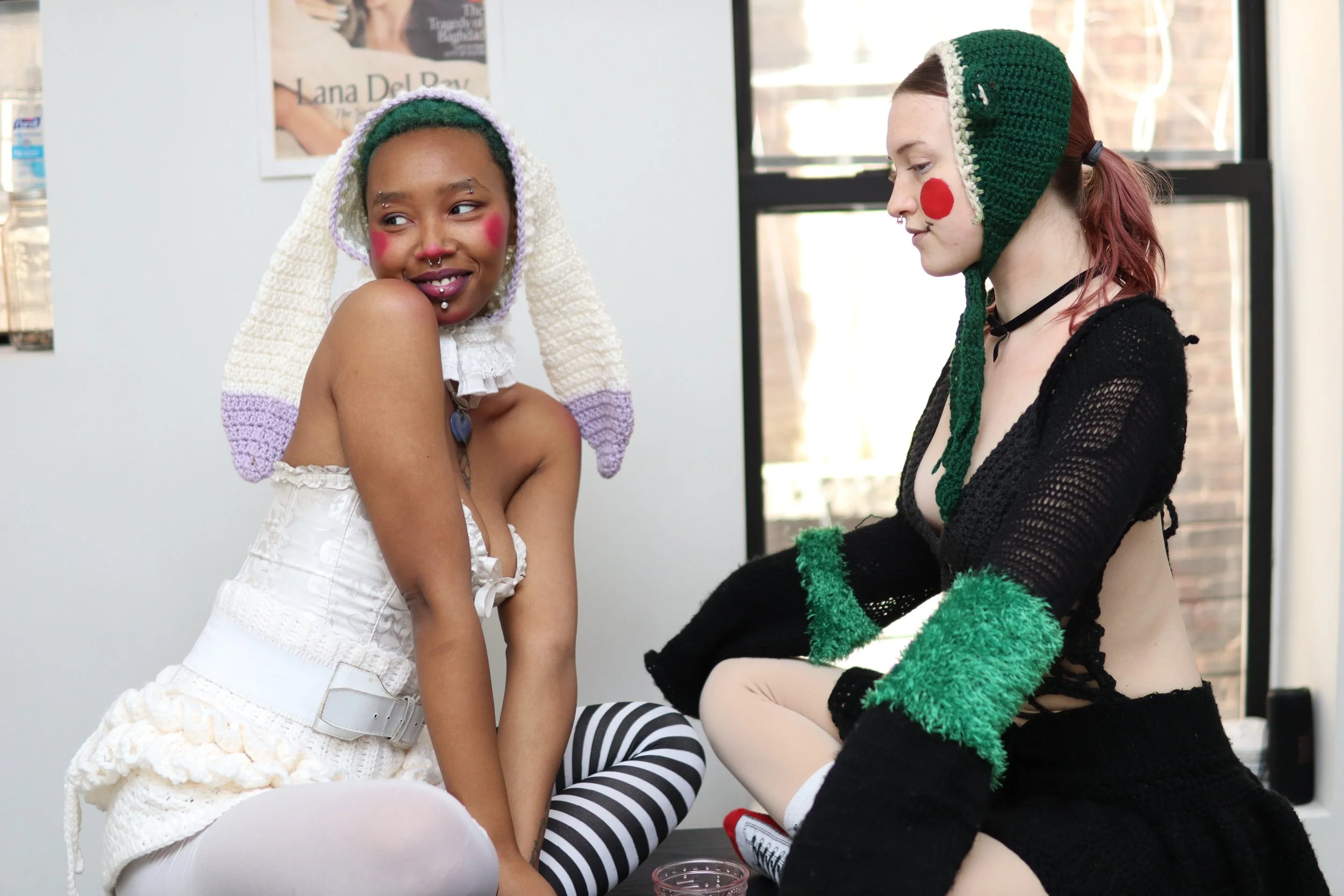 Two young women with colorful makeup and accessories, dressed in playful, eclectic outfits, sitting indoors by a window.