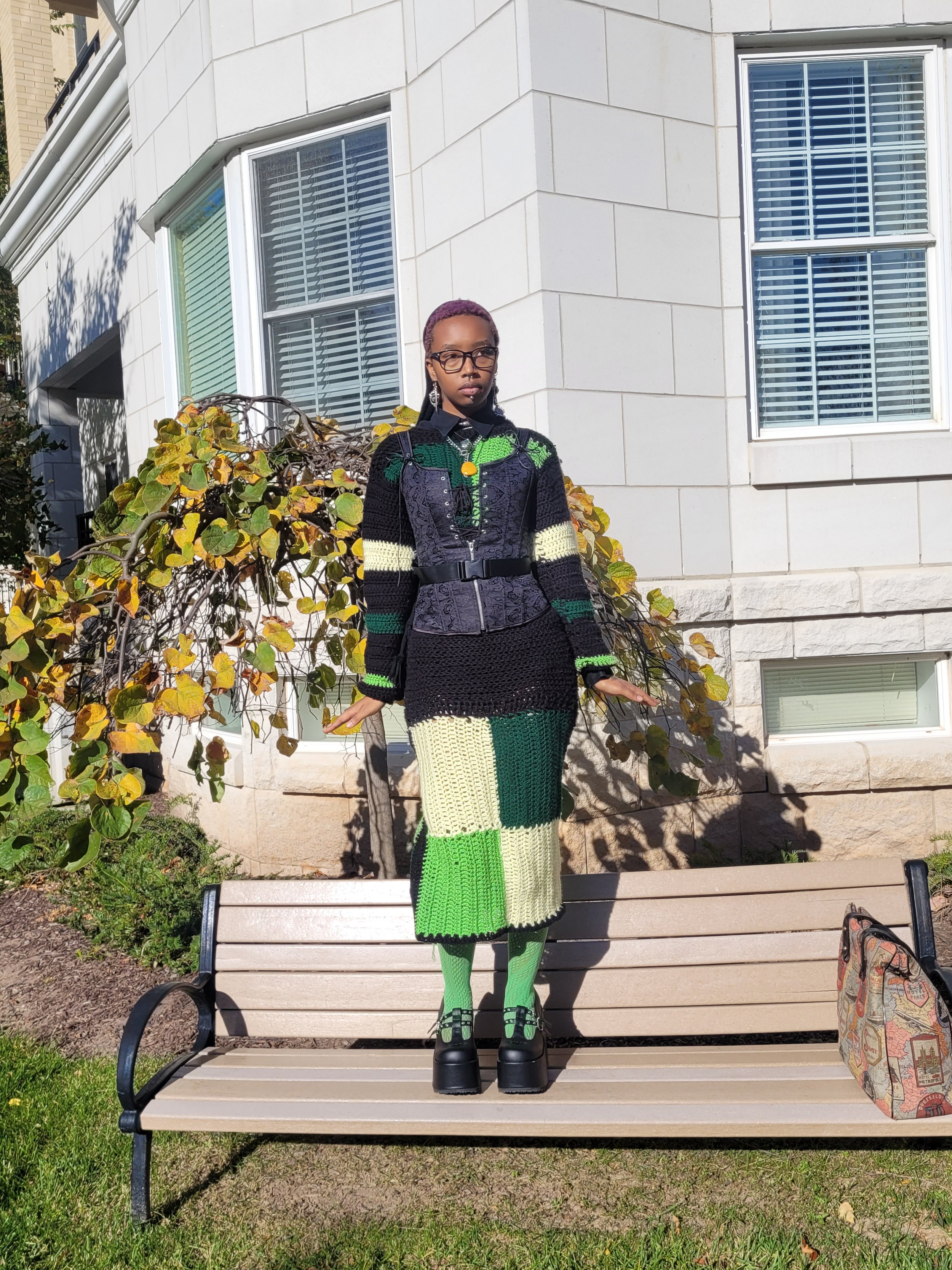 A woman standing on a park bench in front of a white building with large windows, wearing a colorful knitted outfit and green accessories.