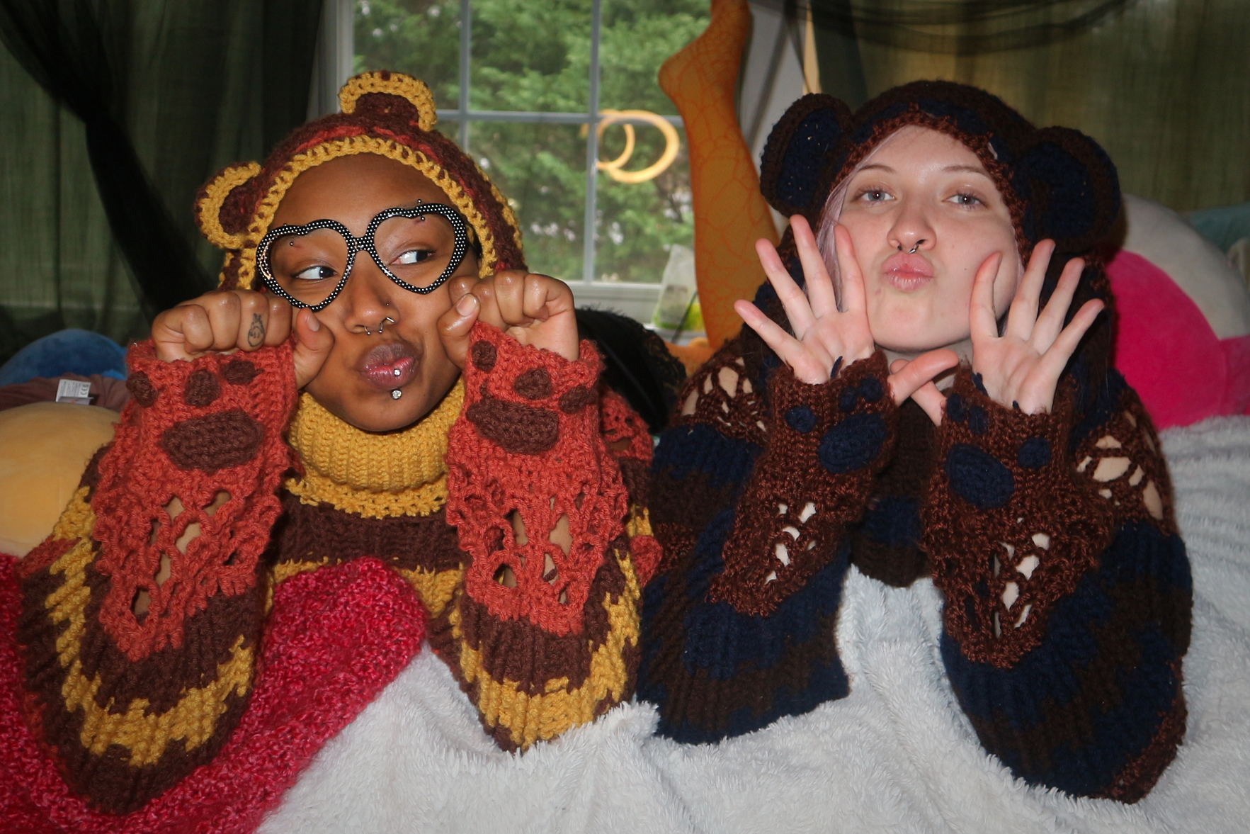 Two young women wearing colorful, crochet bear-themed sweaters and hats posing playfully indoors, with a window and greenery visible in the background.