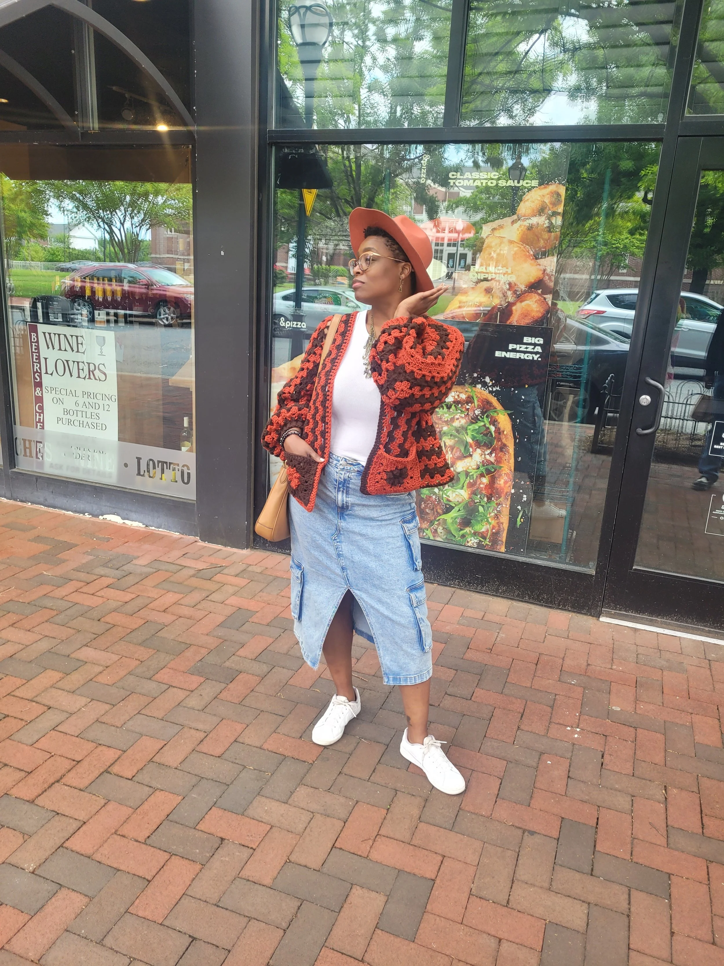 Woman wearing a wide-brimmed hat, glasses, a crocheted jacket, white t-shirt, denim skirt with a slit, and white sneakers, standing outside a restaurant with window posters advertising pizza and tomato sauce.