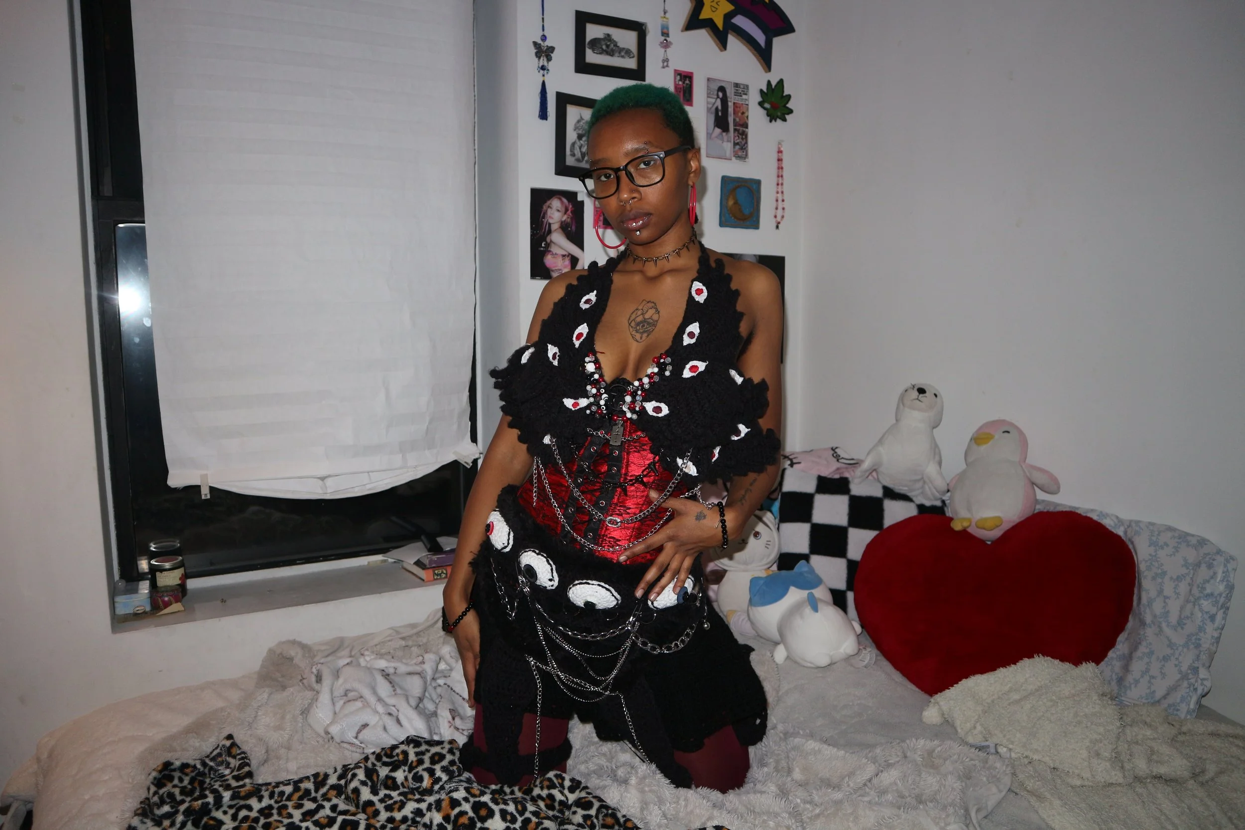 Person with short green hair wearing glasses, a black and red gothic-style outfit with chains and accessories, standing in a bedroom decorated with stuffed animals, plush pillows, and wall art.
