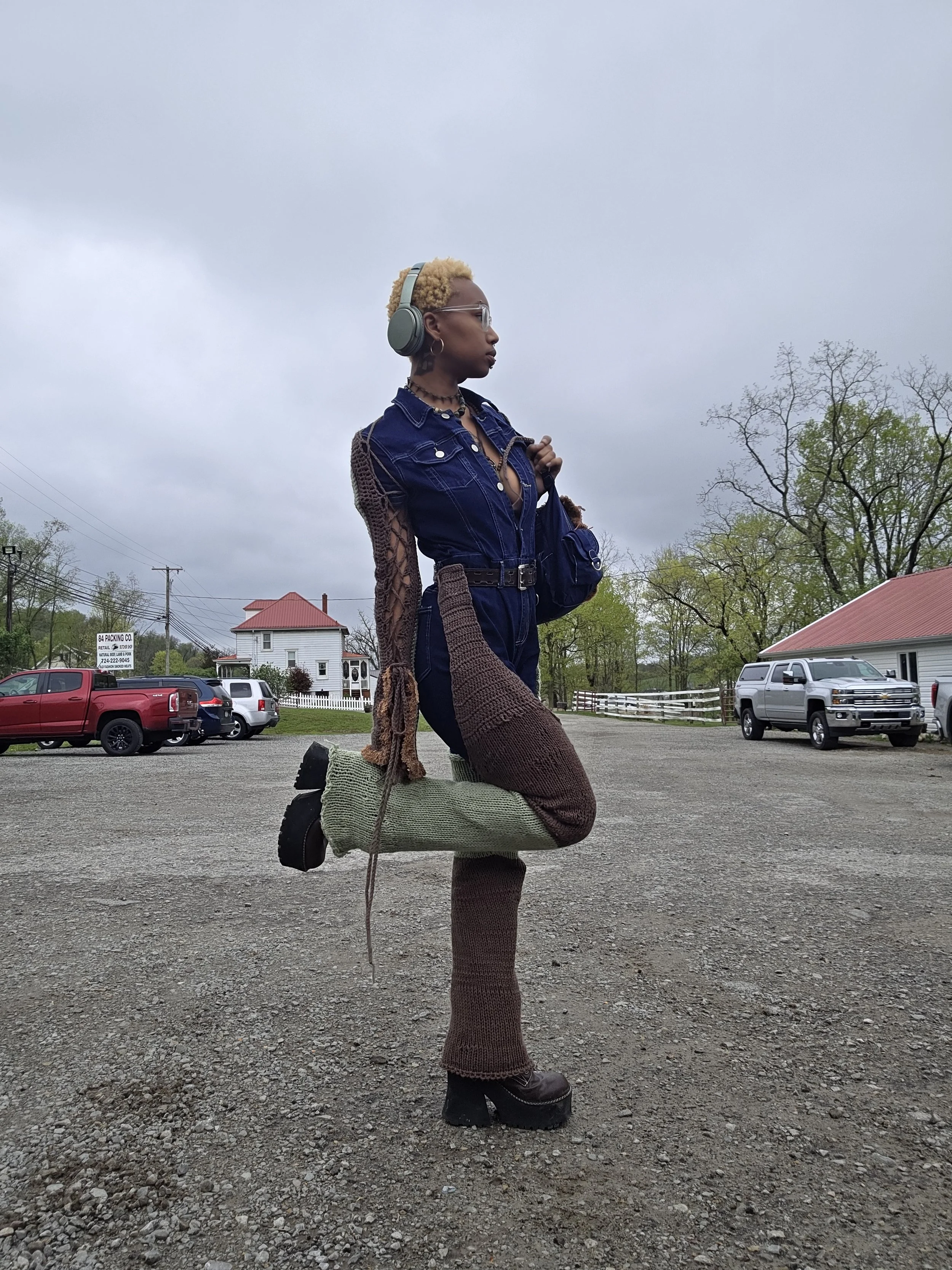 A woman balancing on one leg while holding her other foot, wearing creative boots that resemble knitted socks with a wooden platform heel, on a gravel parking lot under cloudy skies.
